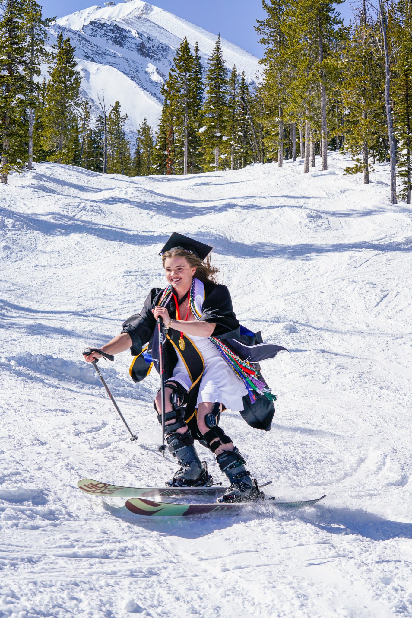 A young woman in a graduation gown and cap skiing down a snowy mountain slope on a sunny day, with trees and a snow-capped mountain in the background.
