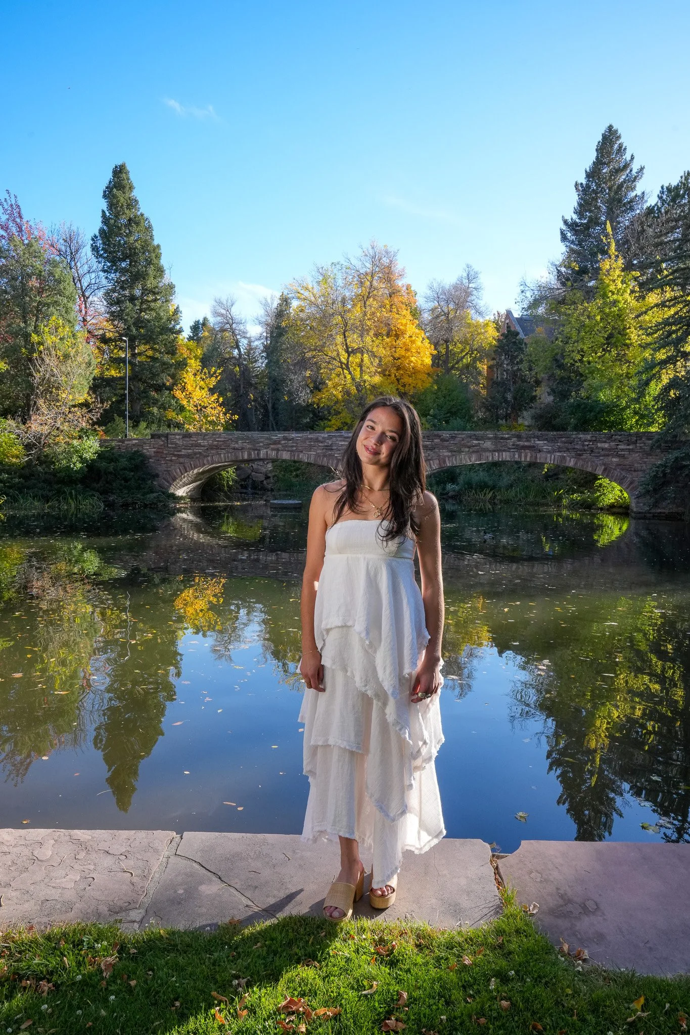 A woman in a white dress standing by a pond with a stone bridge and colorful autumn trees in the background. Graduation photo taken at the University of Colorado Boulder (CU Boulder).