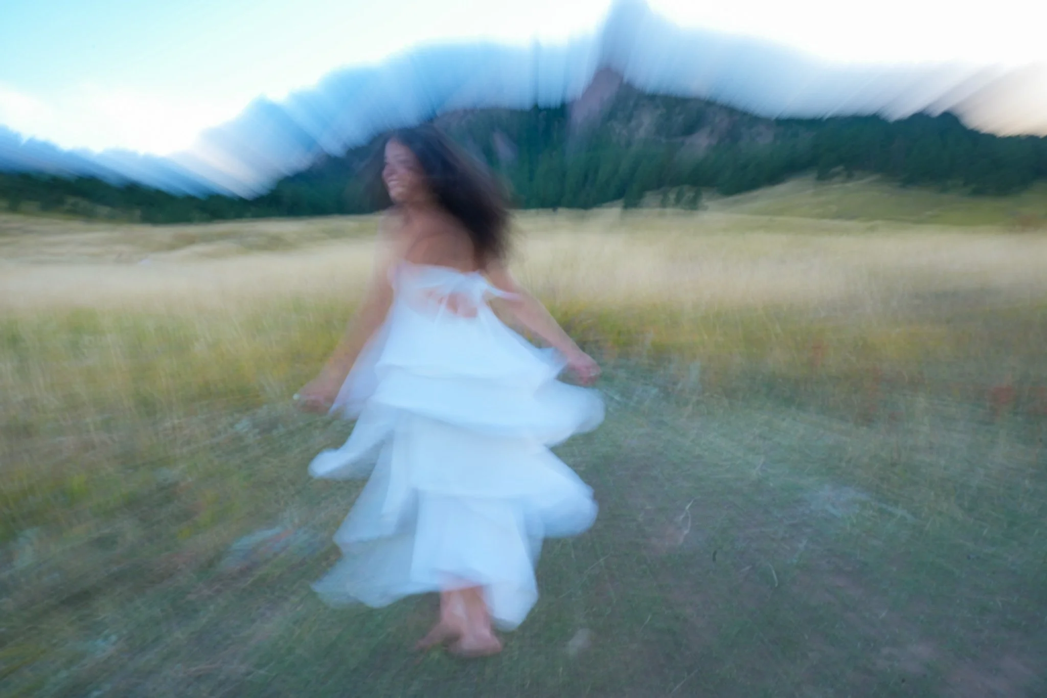 A woman in a white dress dancing outdoors in a grassy field with mountains in the background during daylight. Graduation photo located in front of the Flatiorns at Chautauqua Park.