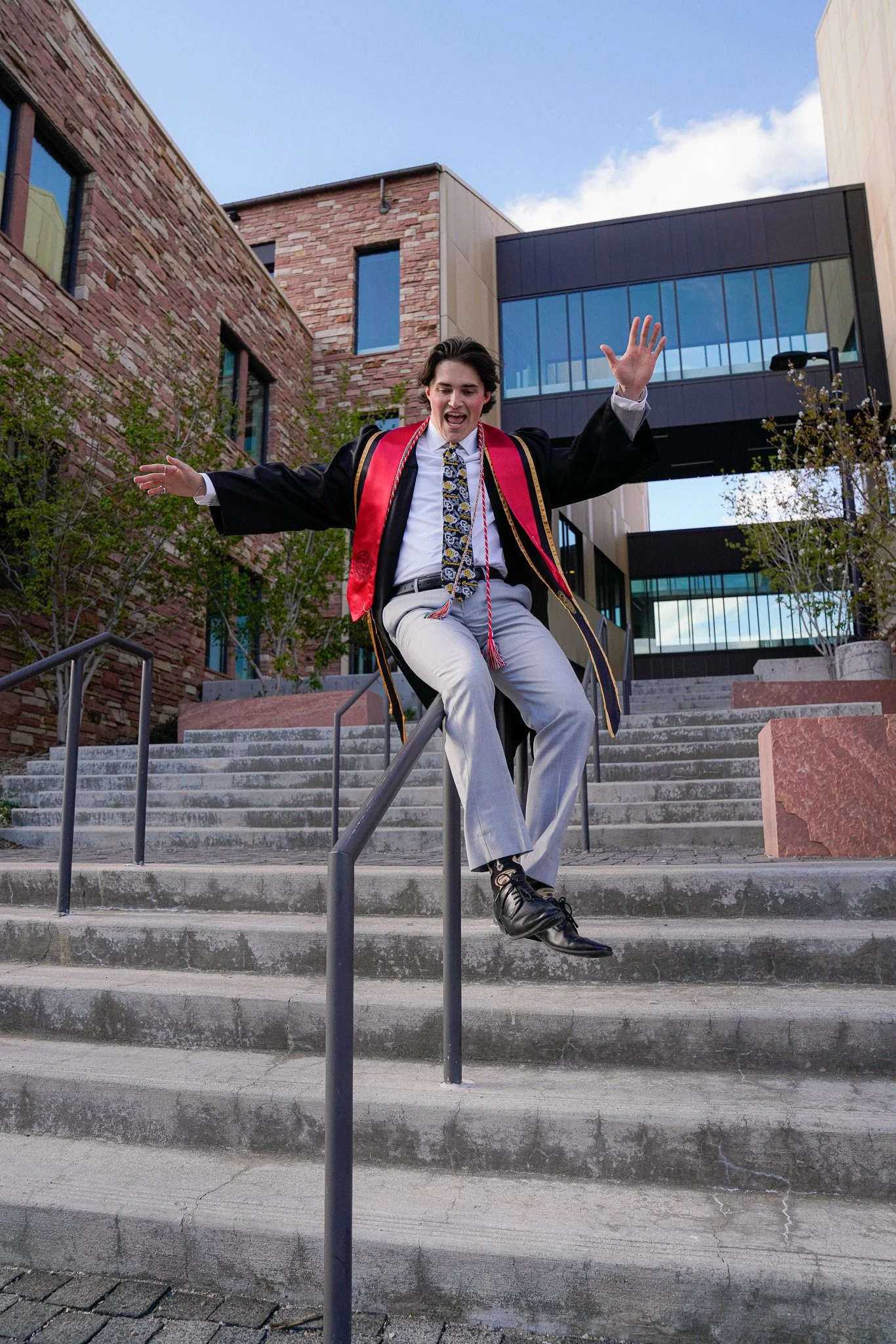 Young man in graduation gown and cap celebrating mid-air on outdoor stone stairs in front of modern university buildings. Graduation photo taken at the University of Colorado Boulder (CU Boulder).