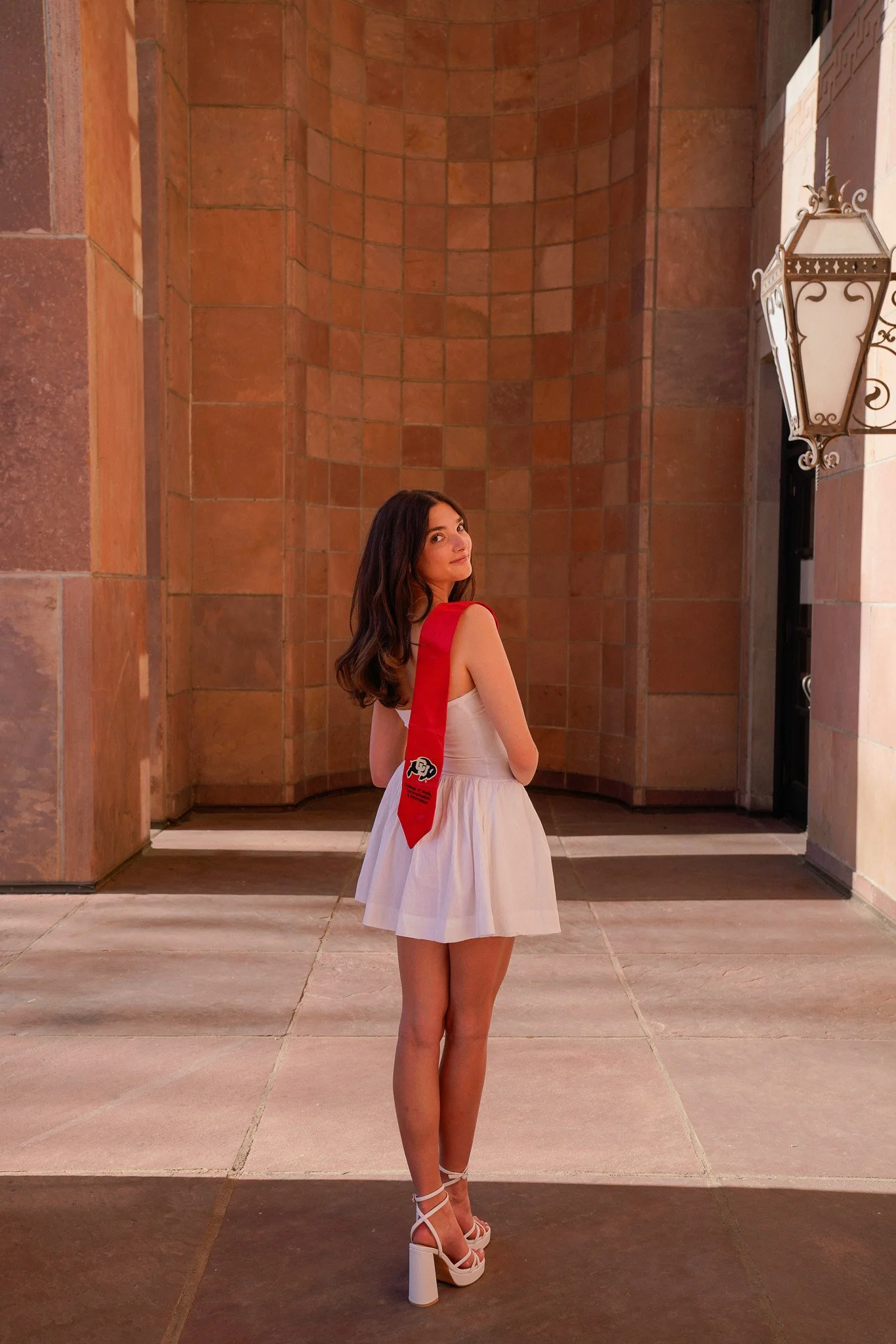 A woman with brown hair wearing a white dress, white high heels, and a red sash standing outdoors near a brick building at night. Graduation photo taken at the University of Colorado Boulder (CU Boulder).