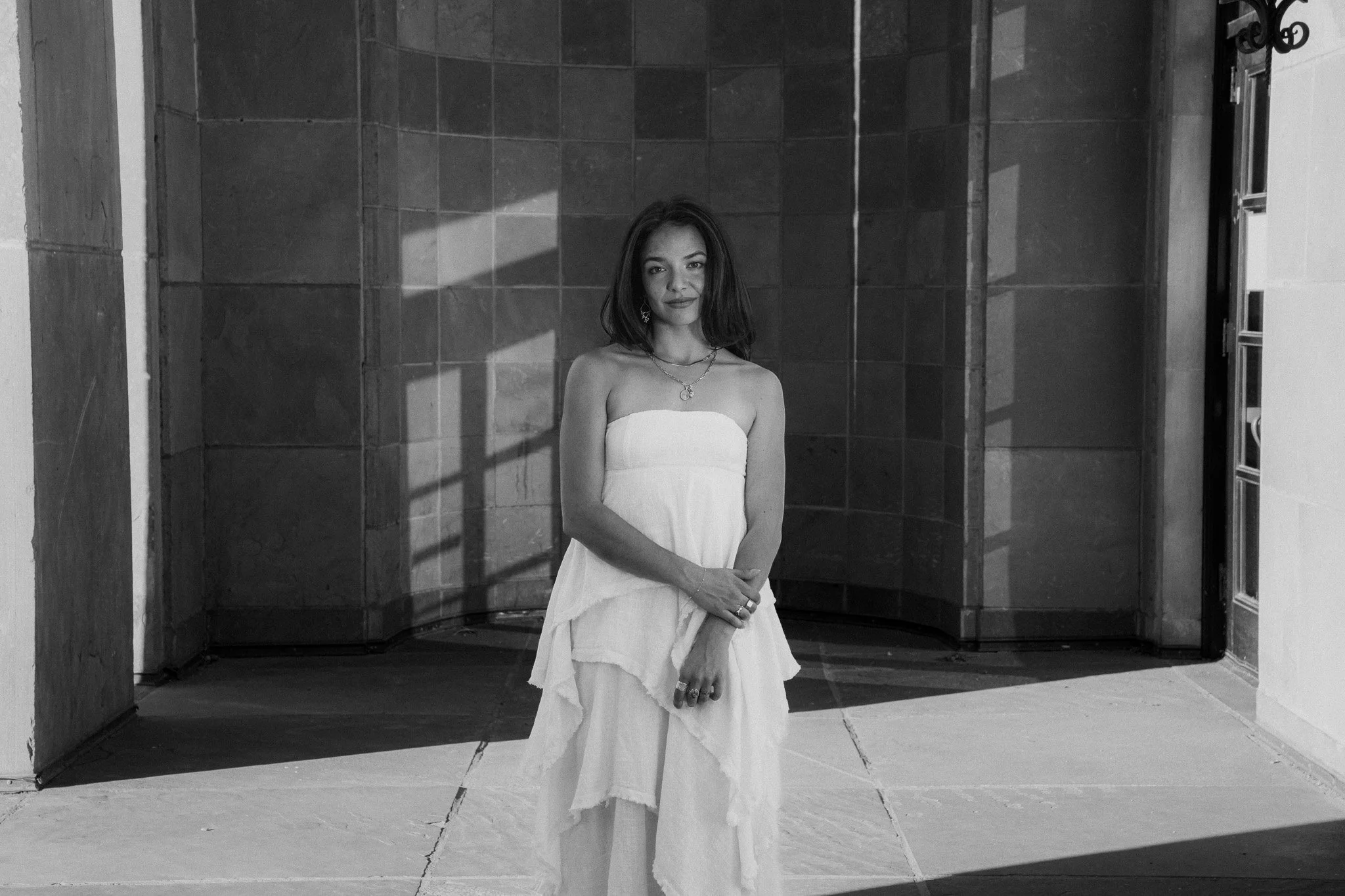 A woman in a white strapless dress standing outdoors with shadows of window panes cast on the wall behind her. Graduation photo taken at the University of Colorado Boulder (CU Boulder).