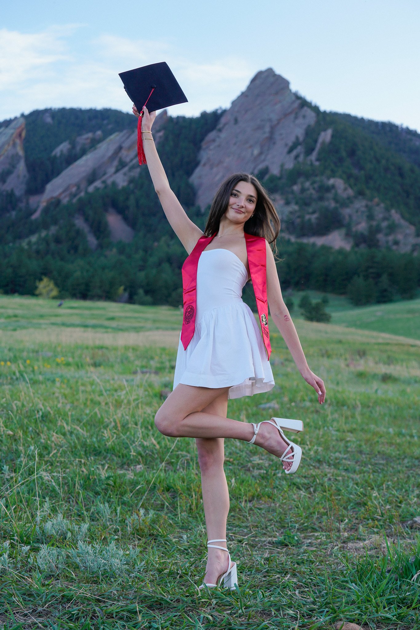 A young woman in a white dress and high heels is celebrating her graduation outdoors, holding a graduation cap in the air, with mountains and a green field in the background. Graduation photo located in front of the Flatiorns at Chautauqua Park.