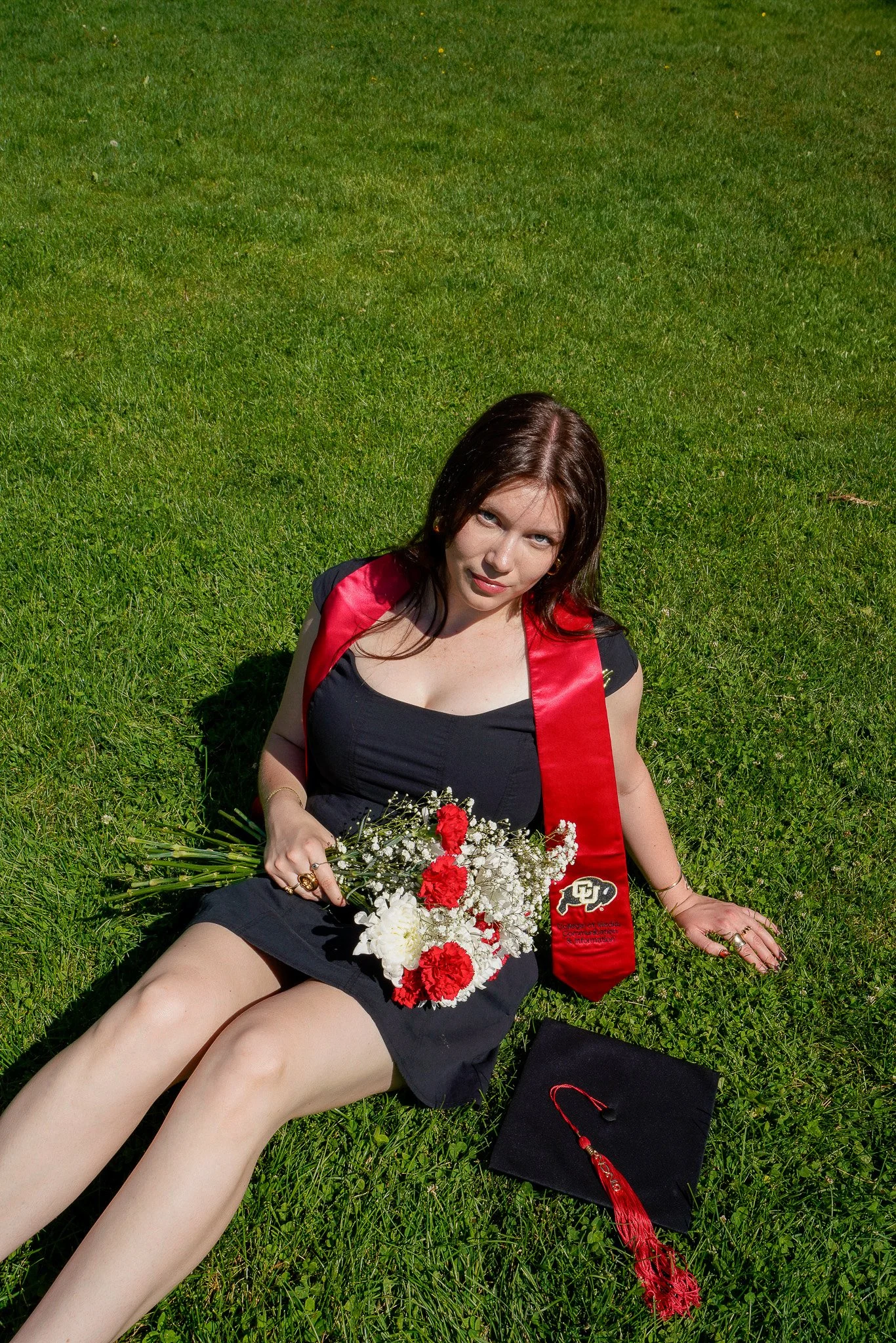 A young woman with brown hair in a black dress and a red graduation stole sitting on green grass, holding a bouquet of red and white flowers, with a black graduation cap and a red tassel nearby.