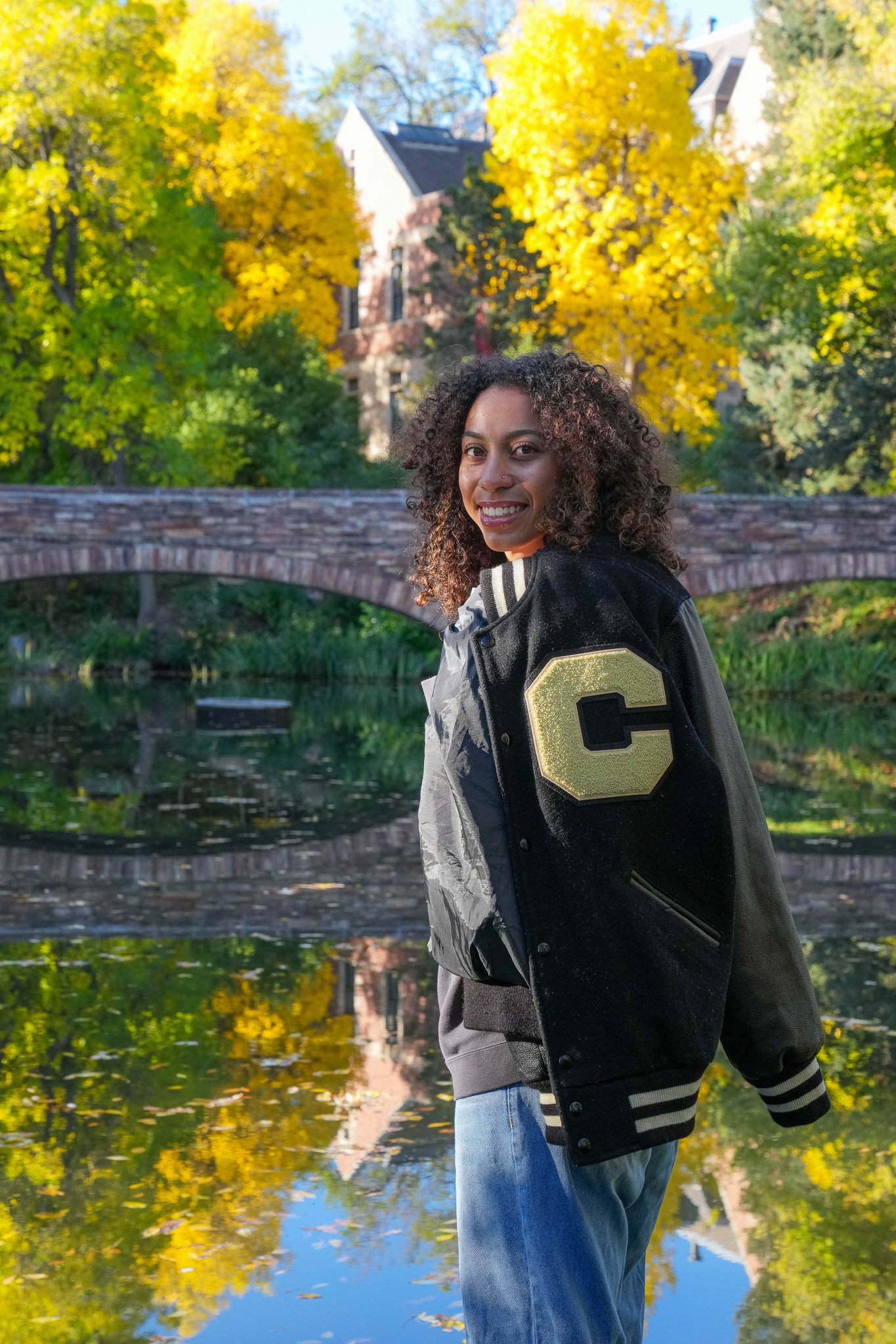 Young woman with curly hair smiling outdoors during fall, wearing a black and gray varsity jacket with a gold letter C, standing near a pond with a stone bridge and colorful autumn trees in the background. Graduation photo taken at the University of 