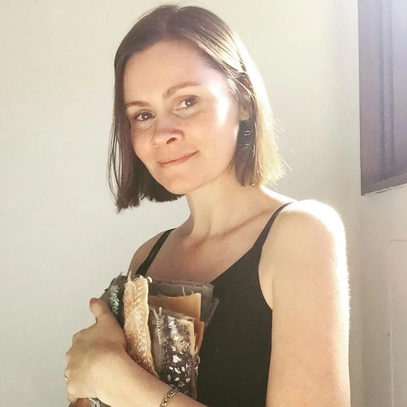 A woman with short brown hair wearing a black spaghetti strap top, holding a stack of various textile artworks, standing in front of a plain light-colored wall with a window visible to the right.