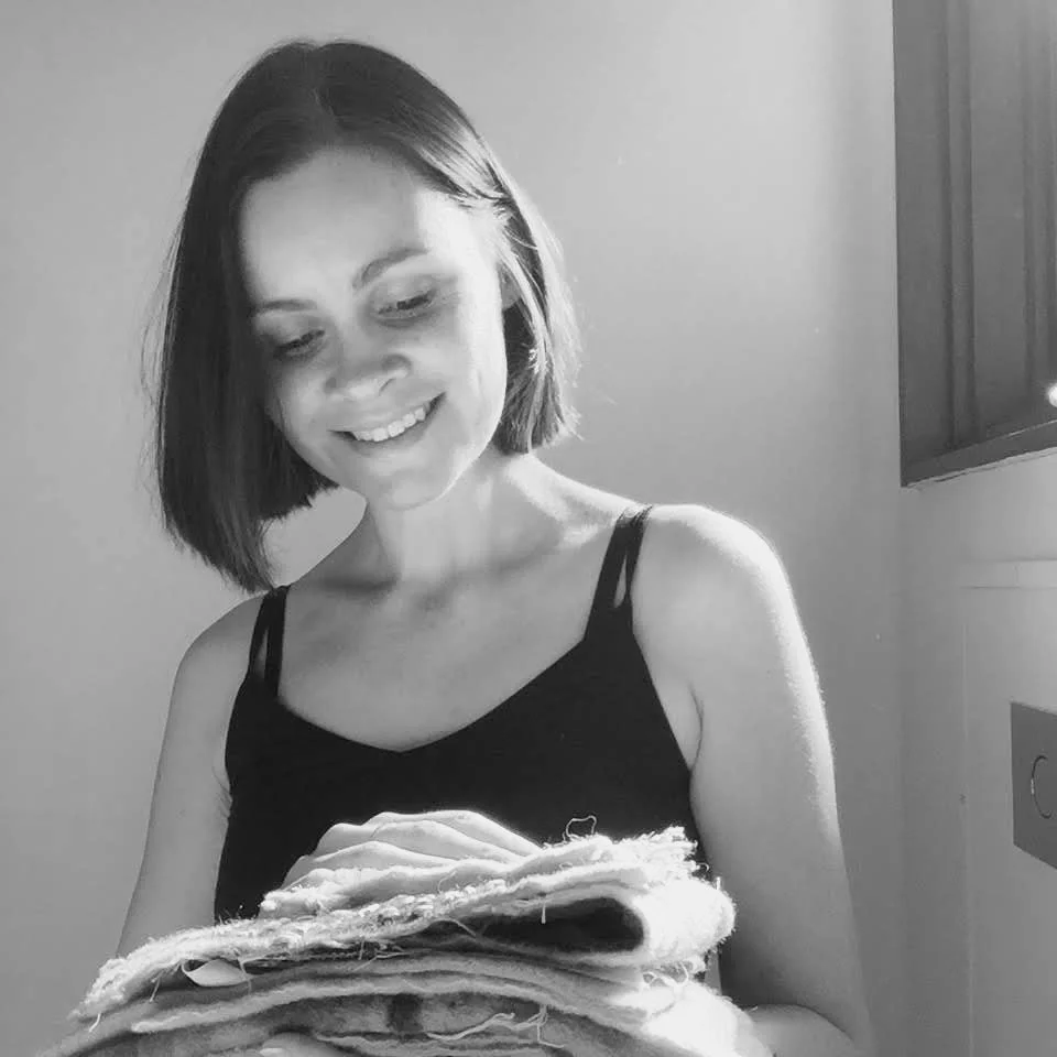 A woman with short hair wearing a black sleeveless top is looking down at textile artworks she is holding, smiling.