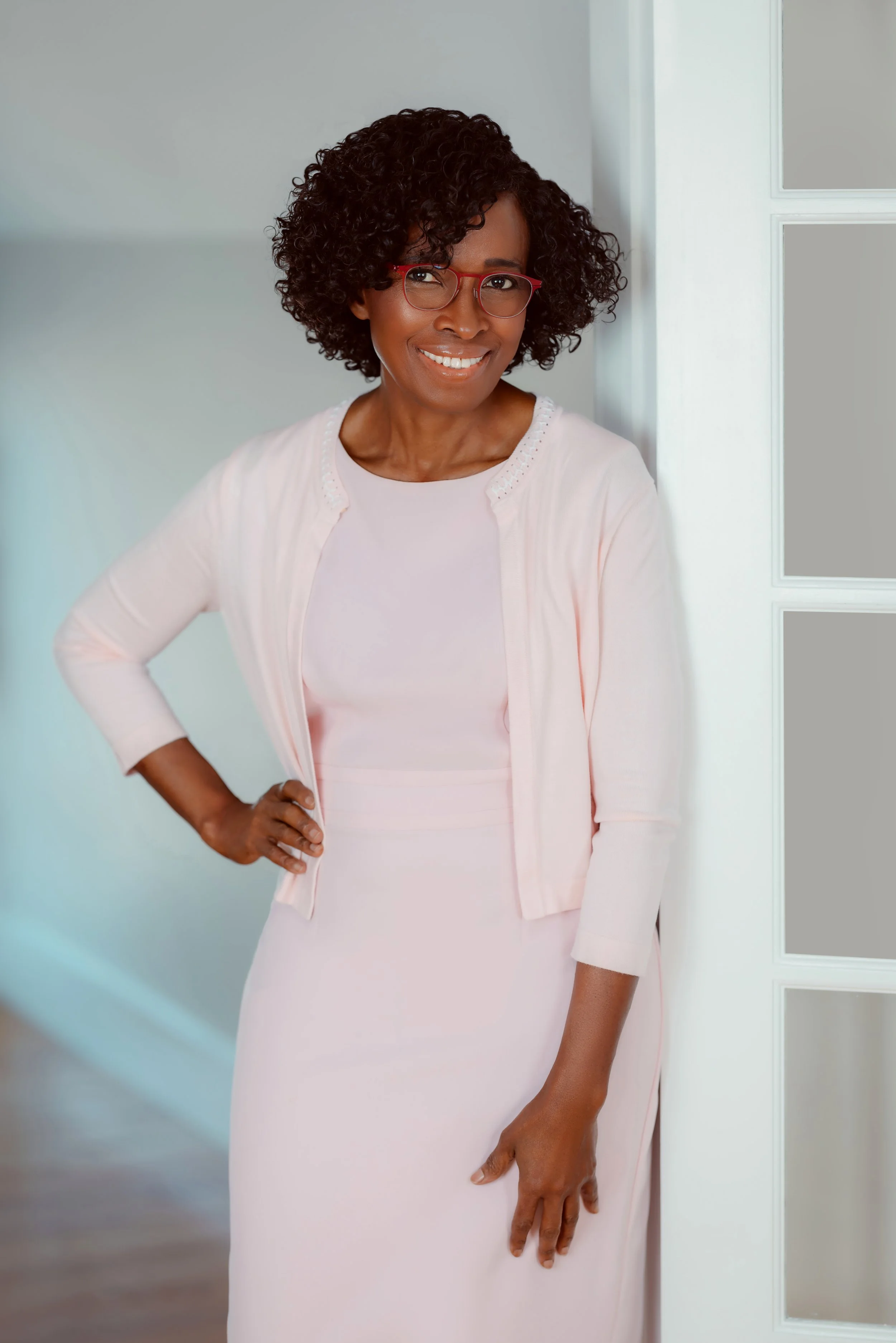 A woman with curly hair and red glasses smiling while standing in a doorway, wearing a light pink dress and matching cardigan.