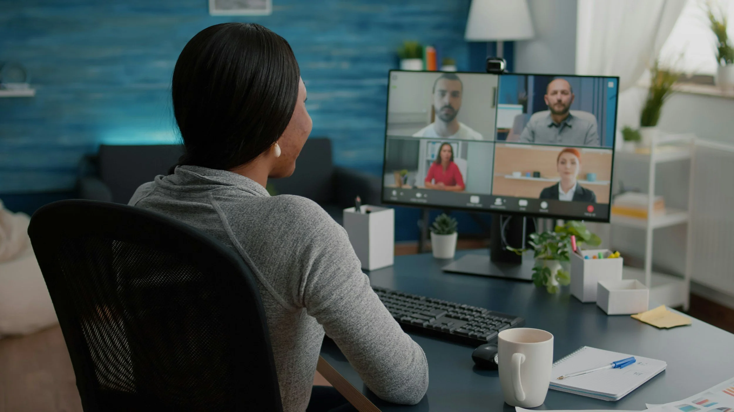 Woman participating in a virtual meeting with four colleagues on her computer monitor, seated at a desk with a coffee mug, notebook, and office supplies.