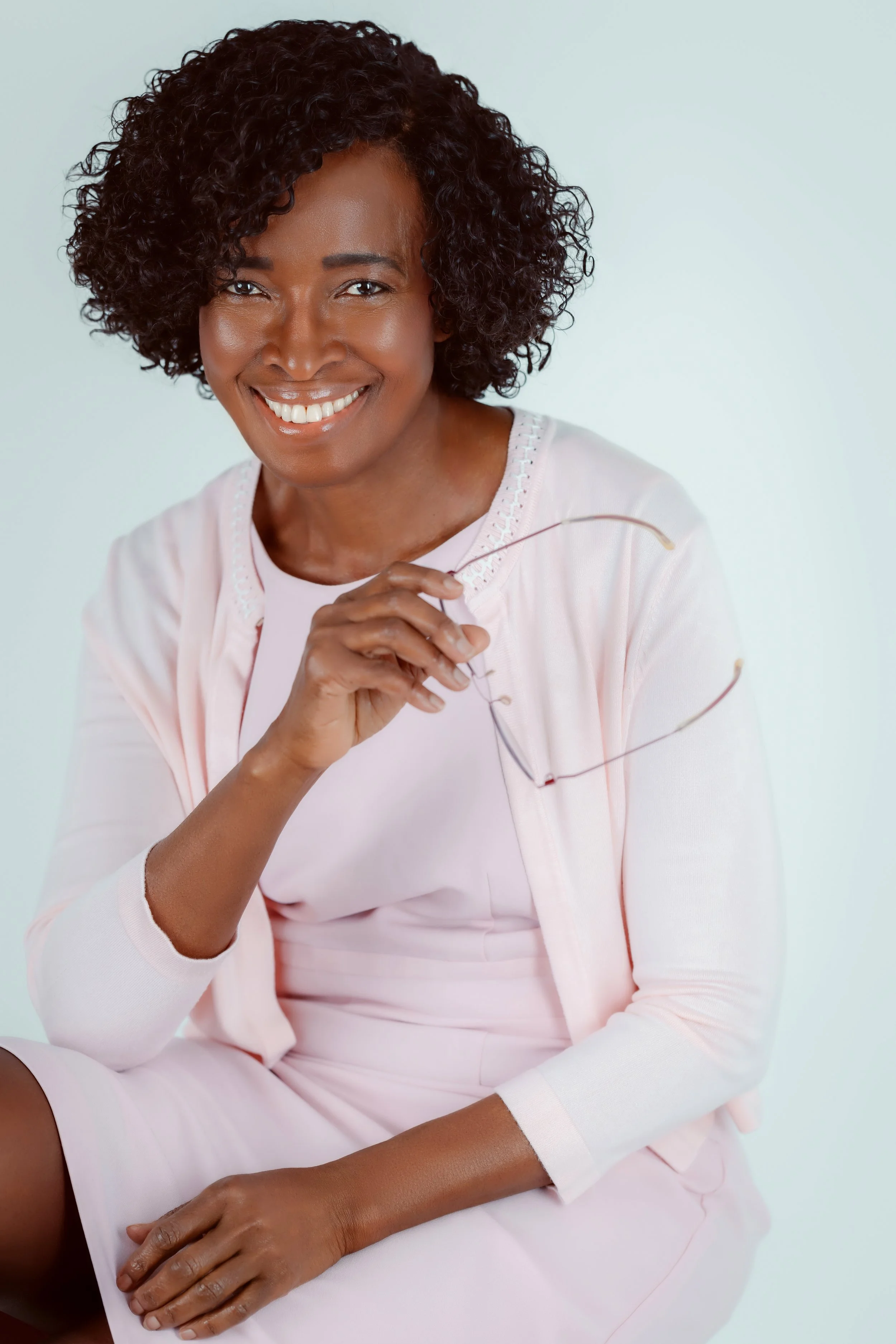 A smiling woman with dark curly hair wearing a light pink dress and jacket, holding eyeglasses, sitting against a plain background.