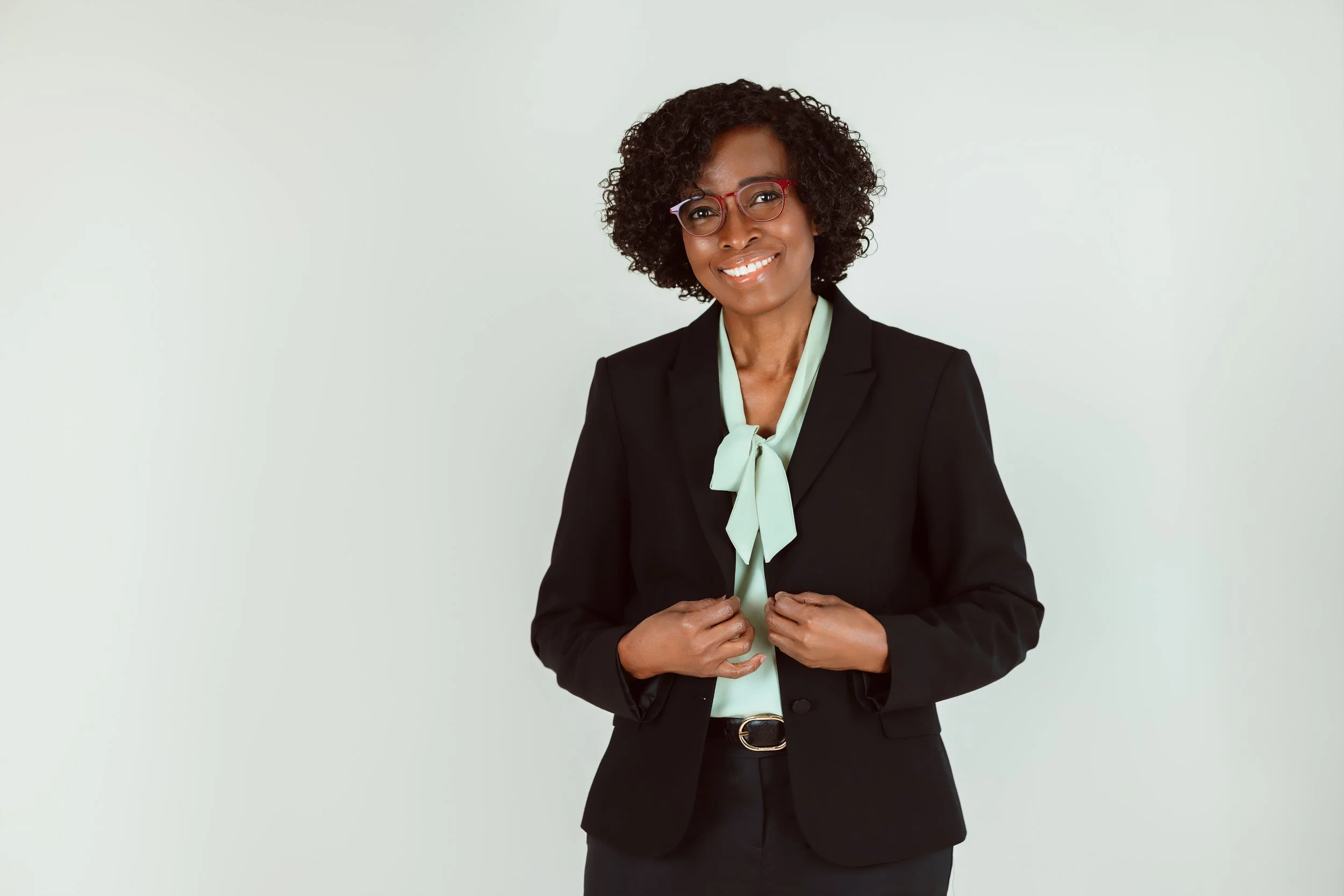 An African American woman with curly hair, wearing glasses, a black blazer, and a light green blouse with a bow, smiling and standing against a plain light background.