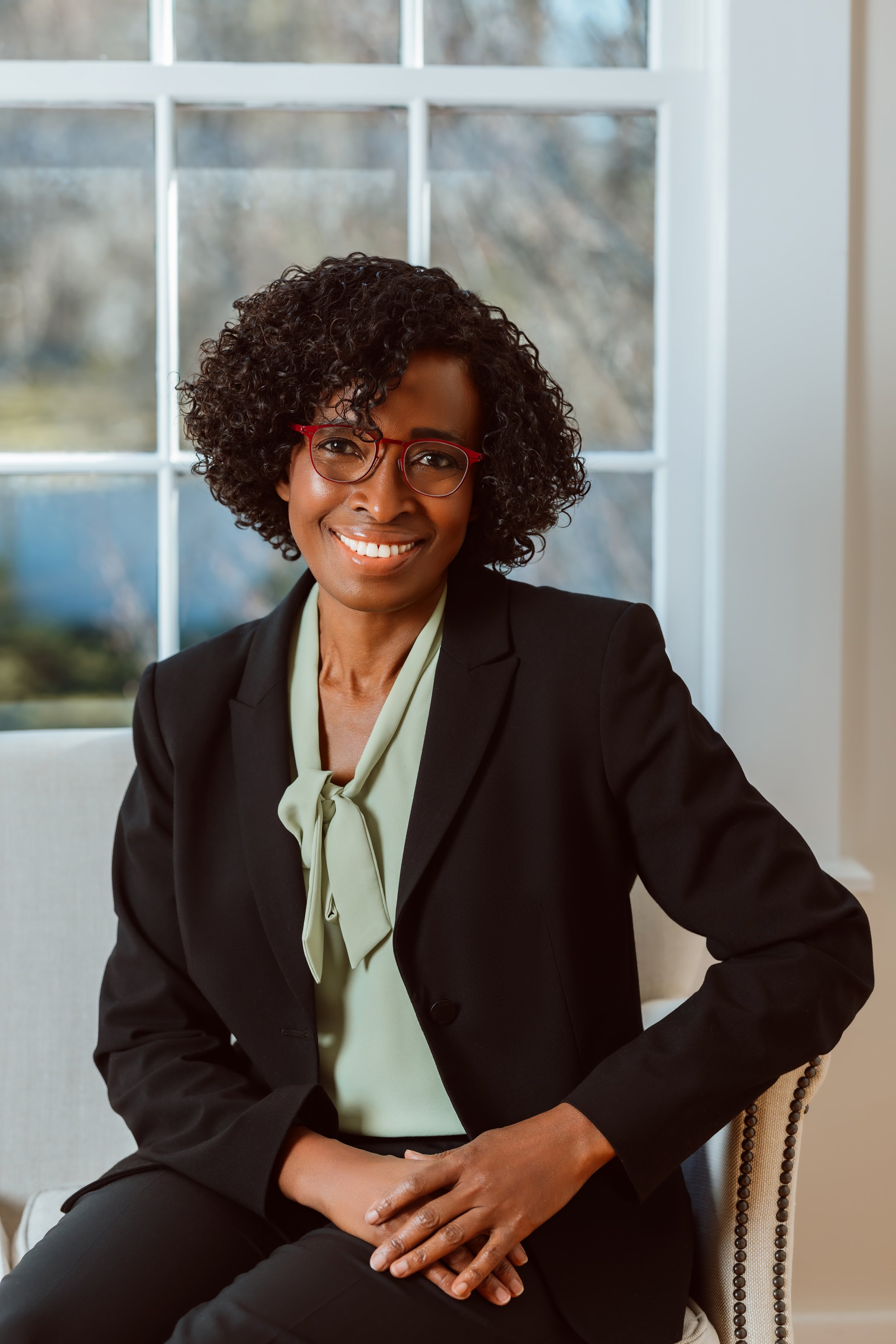 A woman with curly hair and red glasses sitting on a beige chair in front of a large window, wearing a black blazer and light green blouse with a bow.