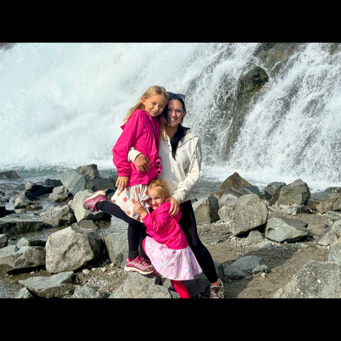 A woman with three young girls on rocks next to a waterfall, all smiling and enjoying their visit.
