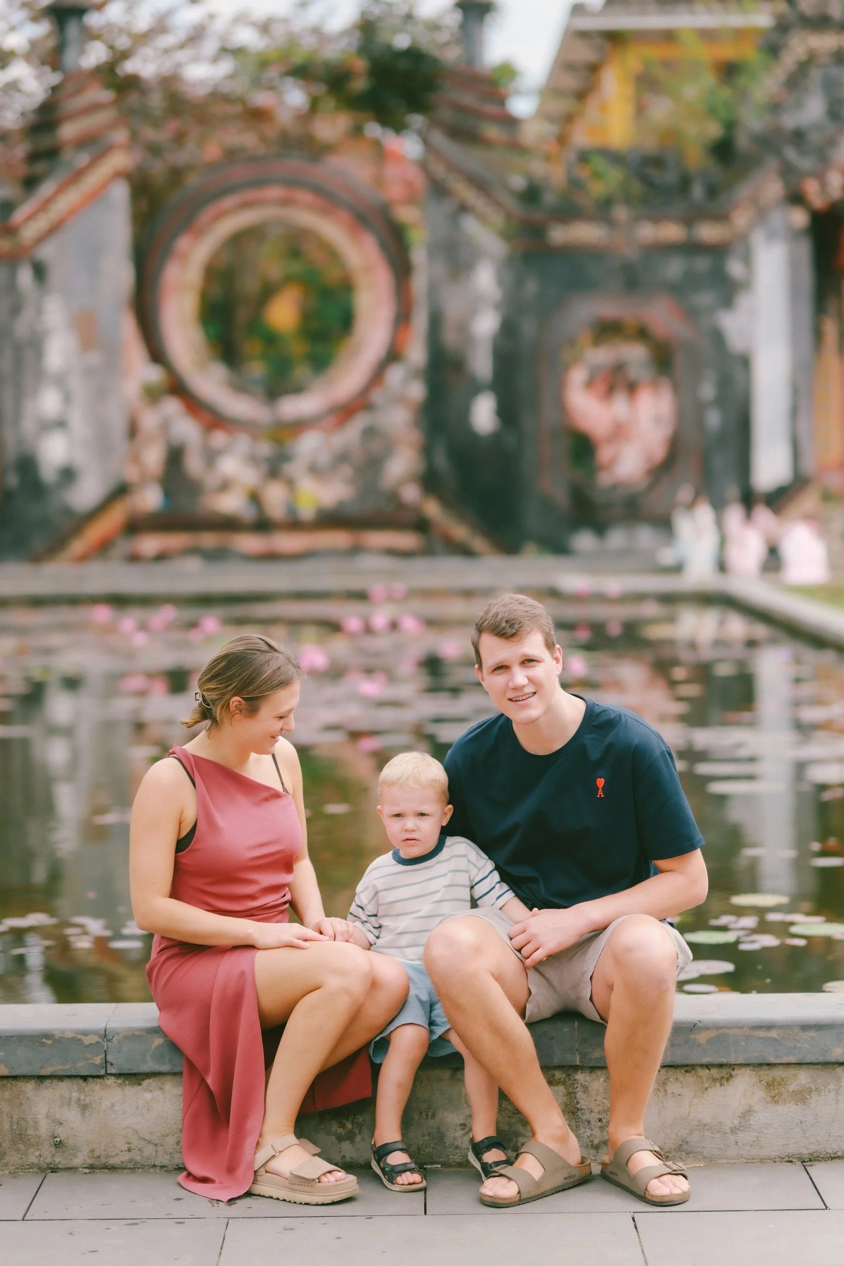 A family of three sitting on a concrete ledge by a water fountain surrounded by pink water lilies, with ornate, colorful architecture in the background.
