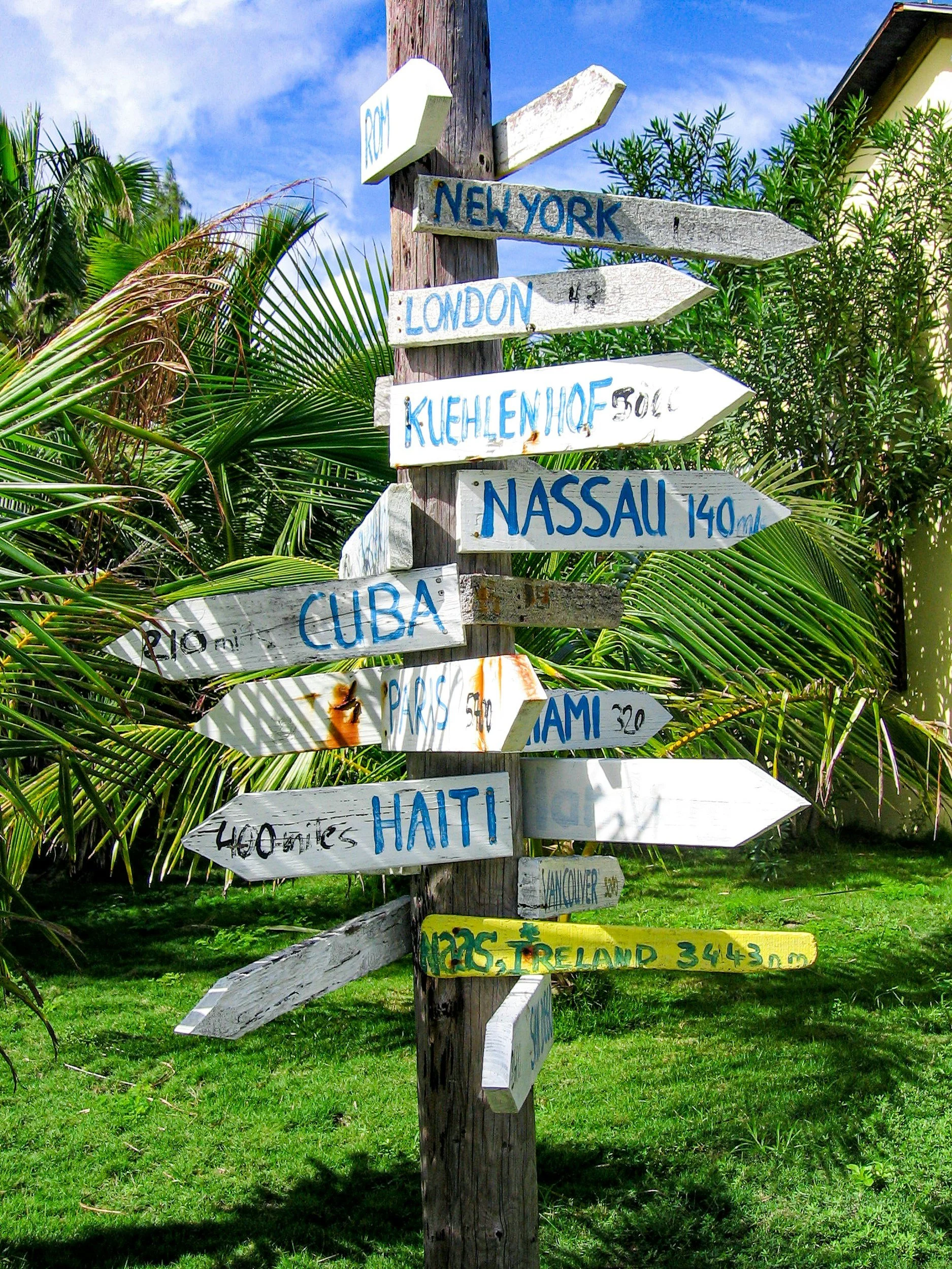 Colorful directional signs on a wooden post pointing to various locations such as New York, London, Kuehlhof, Nassau, Cuba, Paris, Miami, Haiti, Vancouver, and Ireland, with distances marked, set outdoors with lush greenery and a blue sky.