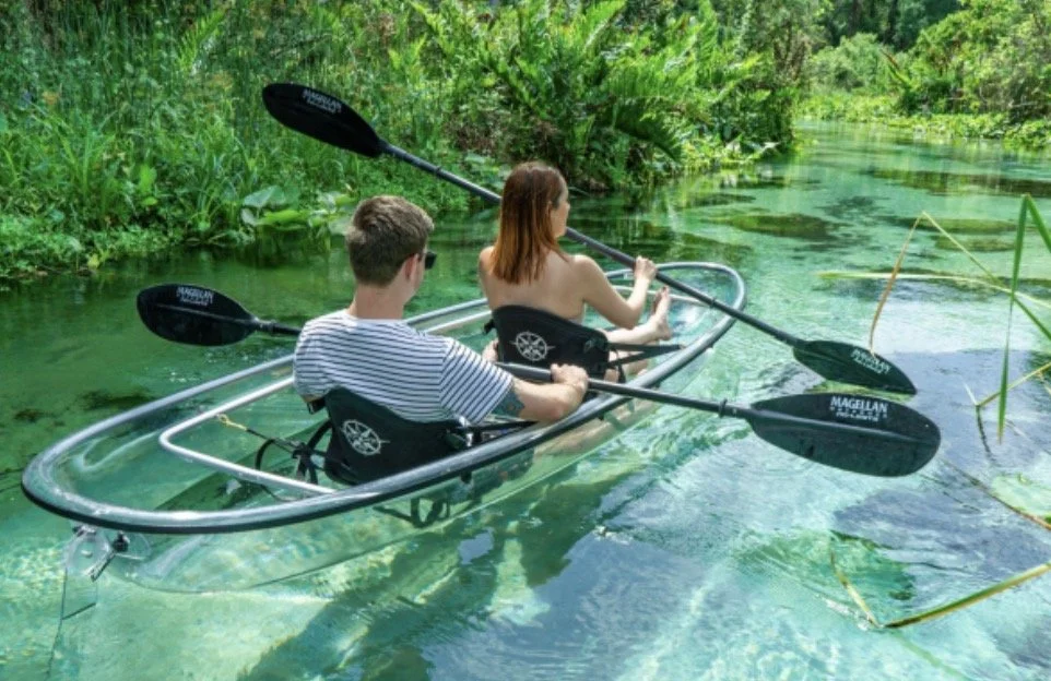 A man and a woman are kayaking on a calm, greenish water canal surrounded by lush, green vegetation.