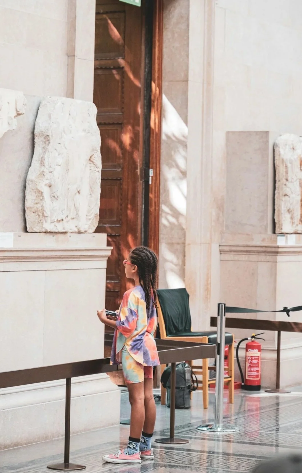 A young girl with braided hair wearing a colorful tie-dye shirt and matching shorts, holding a device, standing in an art museum in front of sculptures and a wooden door.