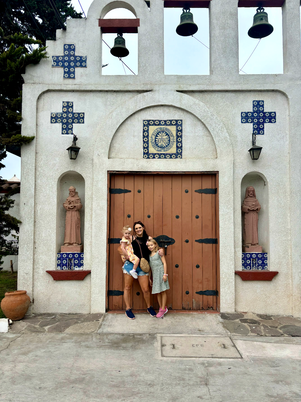 A woman with two young girls standing in front of a church entrance, which features a large wooden door, religious statues, and blue decorative tiles on the white stucco wall.