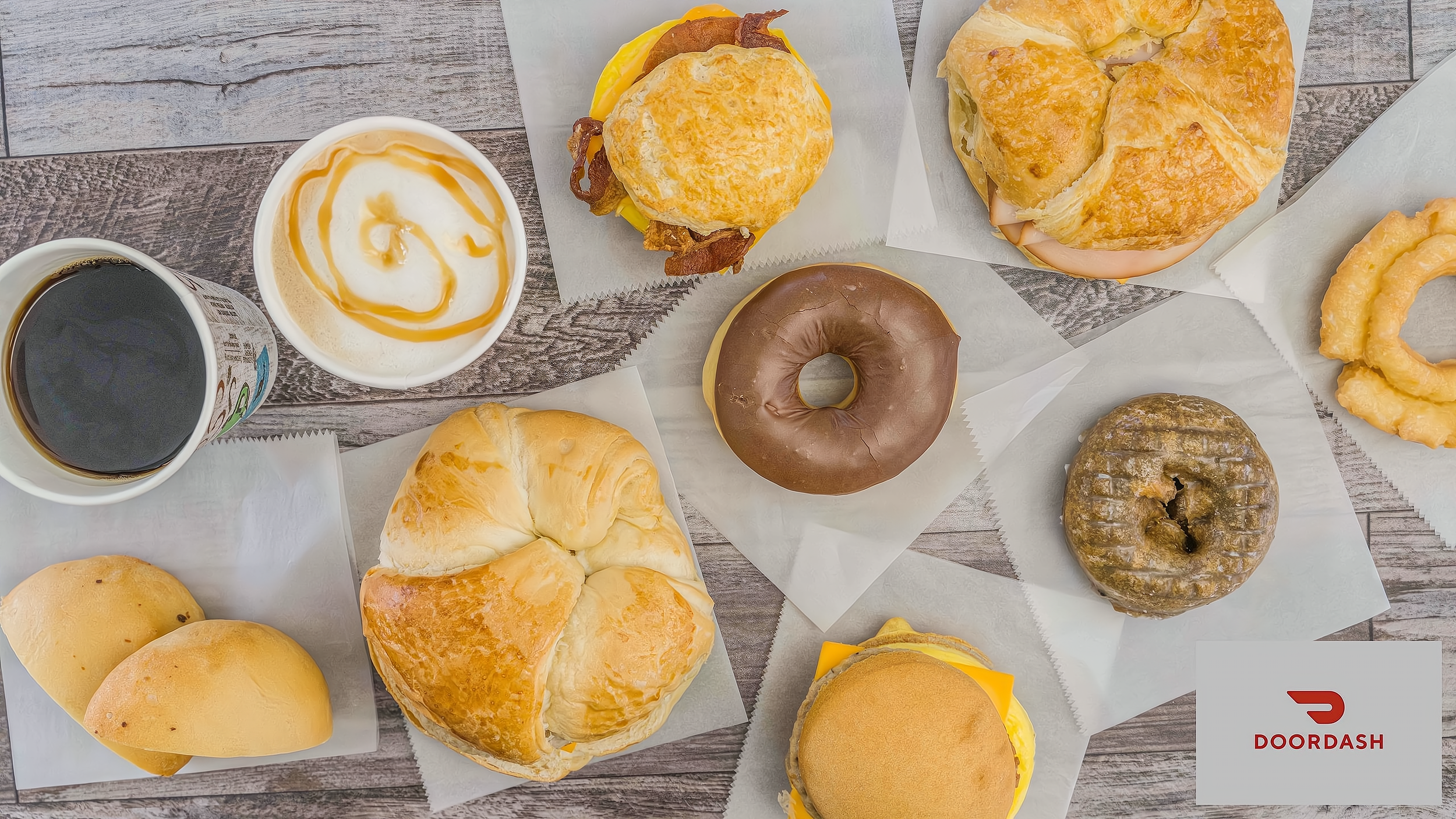 Assorted breakfast items including croissants, muffins, donuts, onion rings, a coffee, and a yogurt with caramel drizzle on a wooden table.