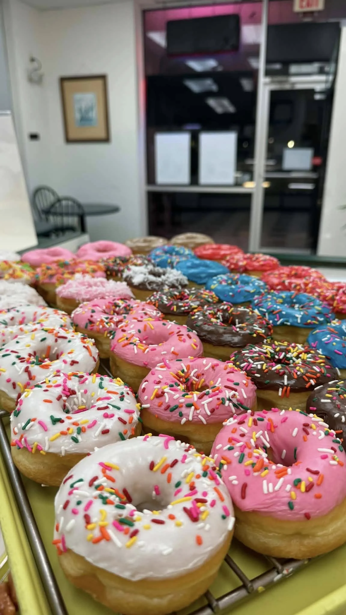 Assorted donuts with colorful sprinkles on a tray inside a store or cafe.