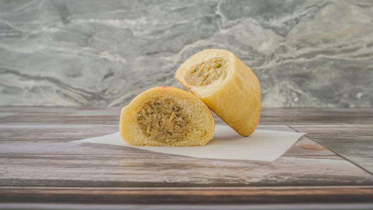 Close-up of a bread roll with a savory filling, cut in half and placed on a piece of white paper on a wooden surface, with a gray marble background.