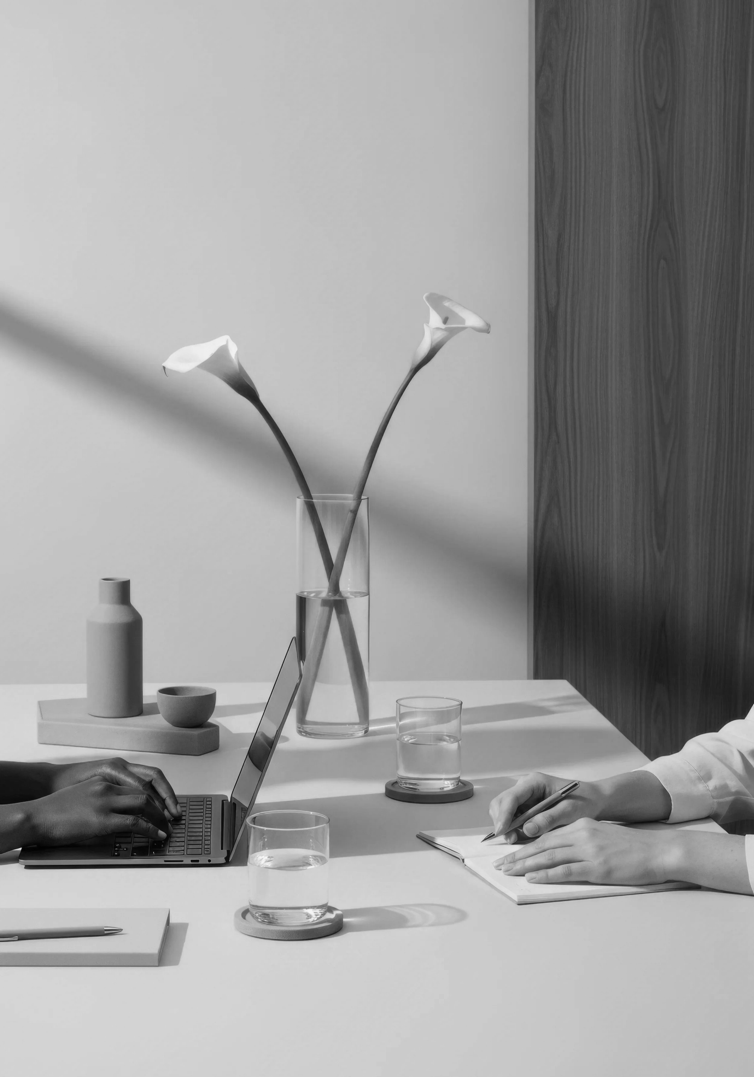 Black and white photograph of a desk scene with two people working; one with dark skin typing on a laptop, the other with light skin writing in a notebook; on the desk are two glasses of water on coasters, a small vase with two calla lilies, a ceramic bottle, a small bowl, and a closed book with a pen on top.