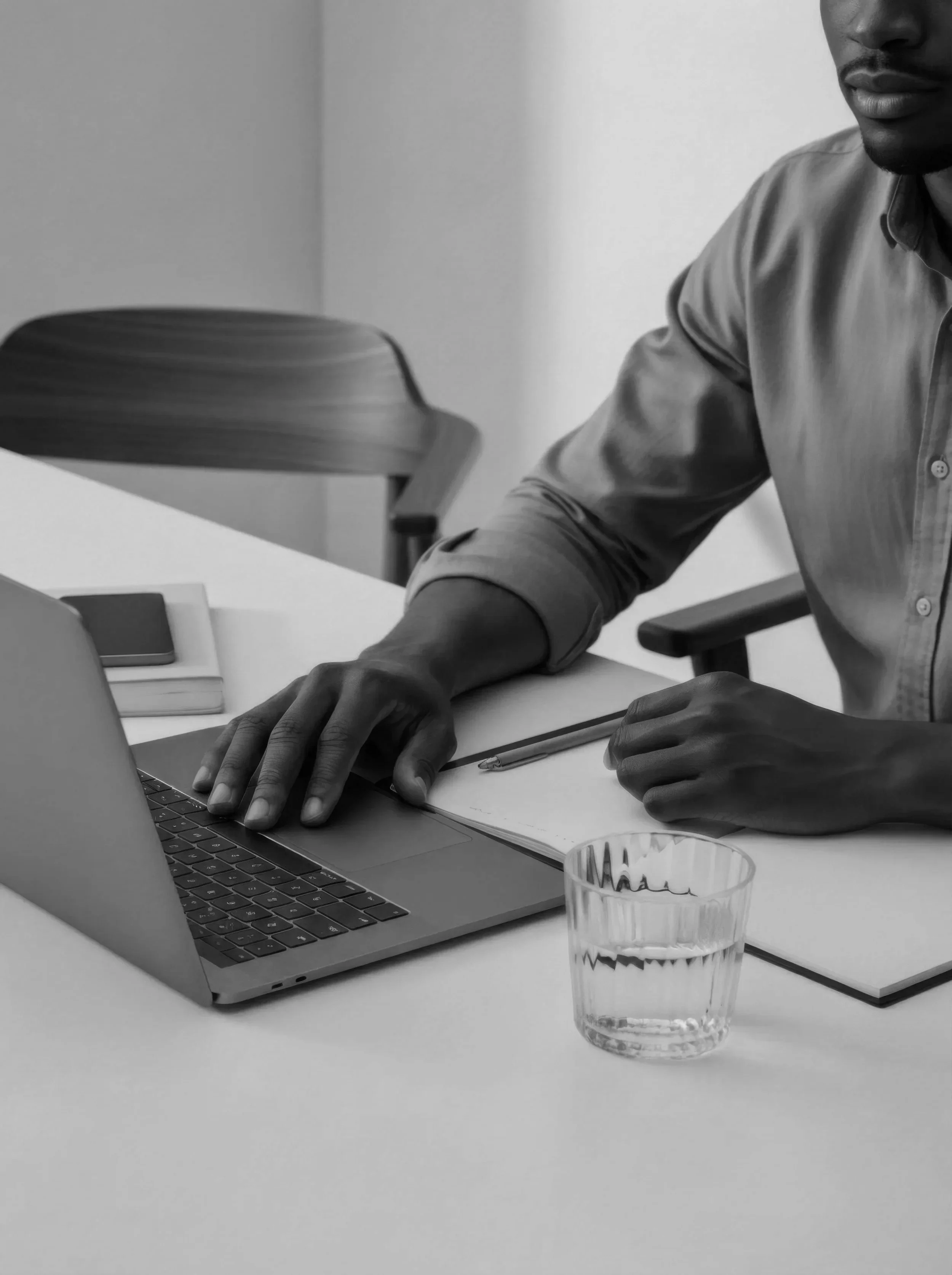 A person wearing a button-up shirt sitting at a desk working on a laptop, with a glass of water, notebooks, and a pen on the desk in black-and-white photograph.