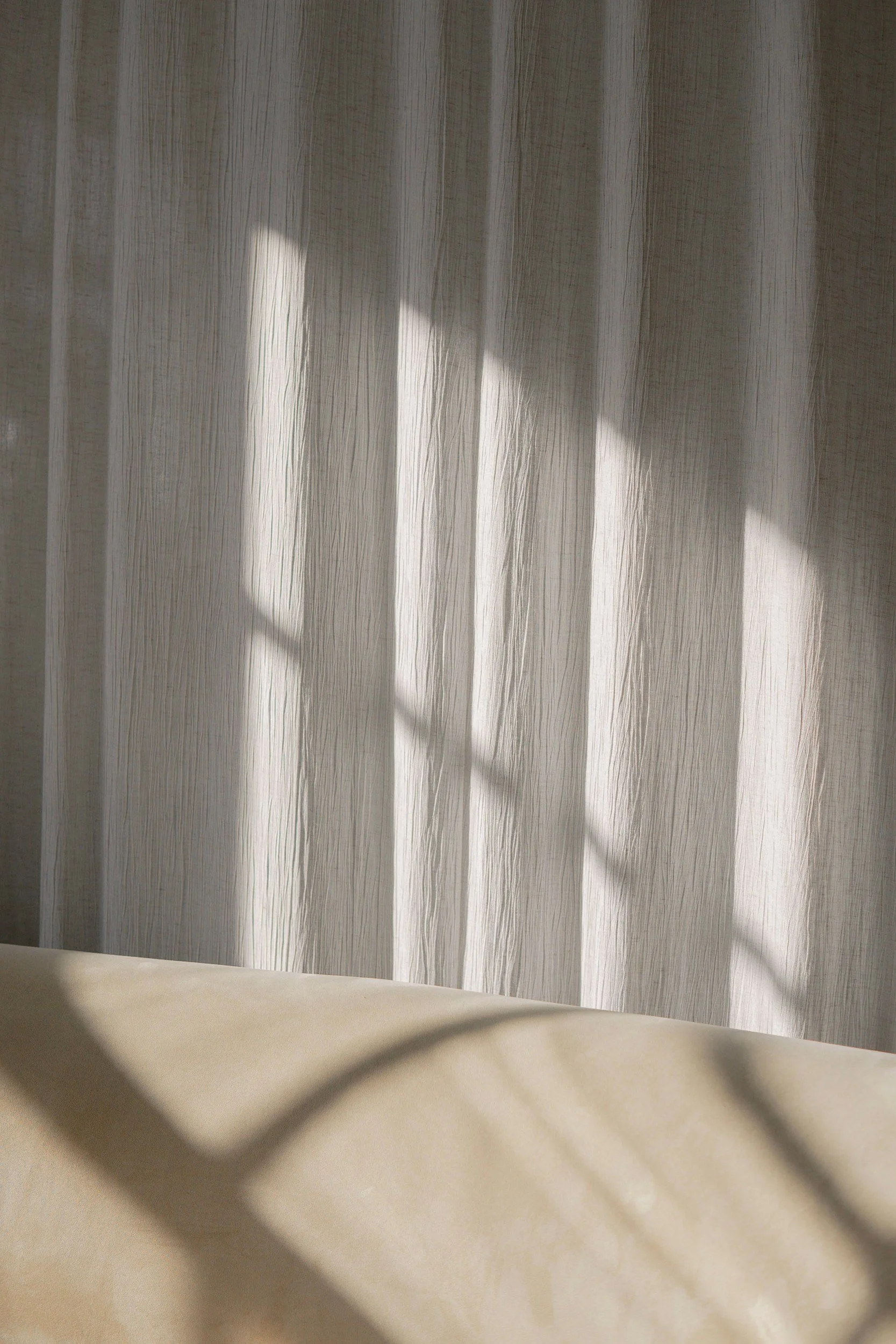 Sunlight casting shadows through a window onto a beige surface and light-colored textured curtains.