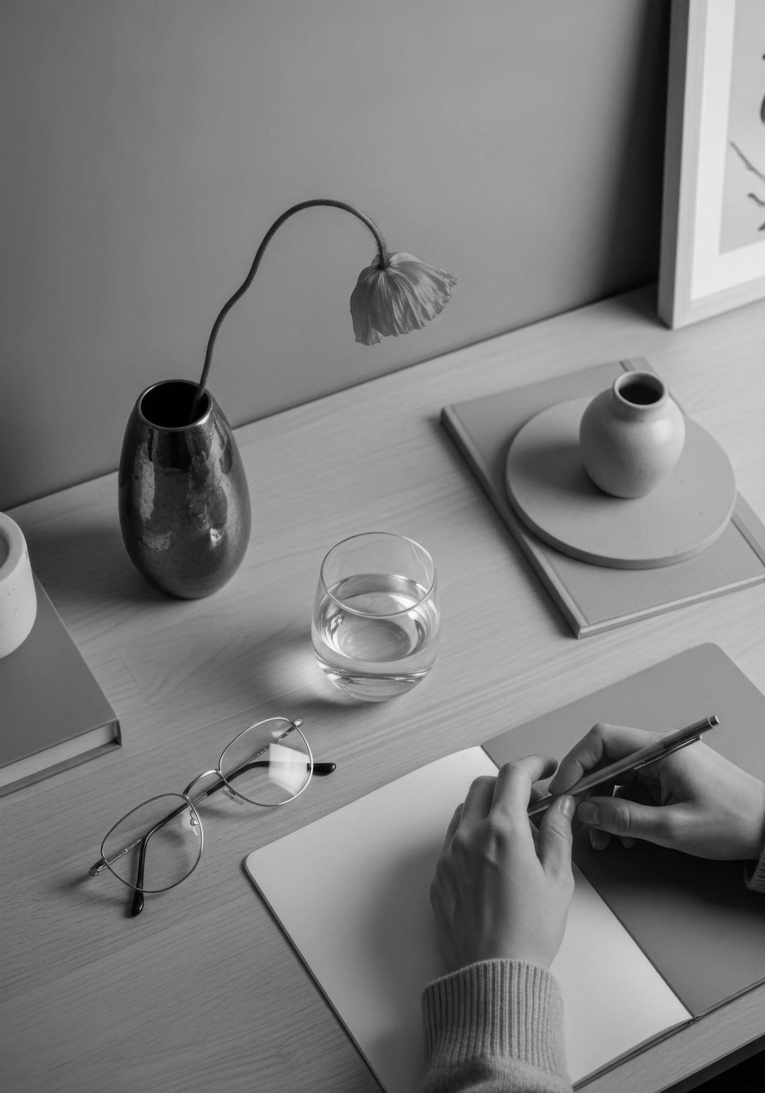 A desk with a vase holding a wilted flower, a glass of water, a pair of glasses, a notepad, and a person's hands writing on a notepad, in black and white.