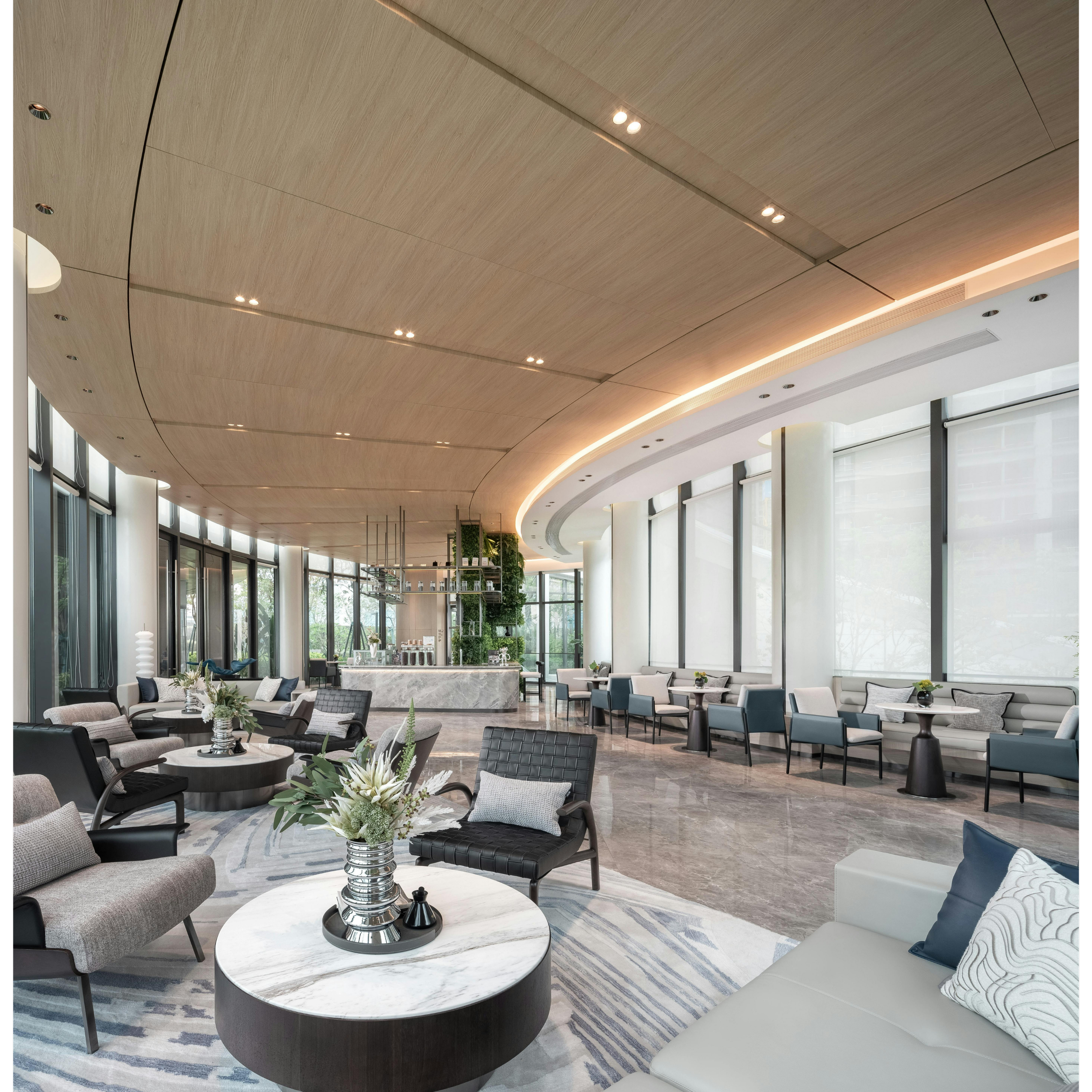 Modern lobby or lounge area with large windows, a curved wooden ceiling with spotlights, and seating including armchairs, sofas, and round tables with floral centerpieces.