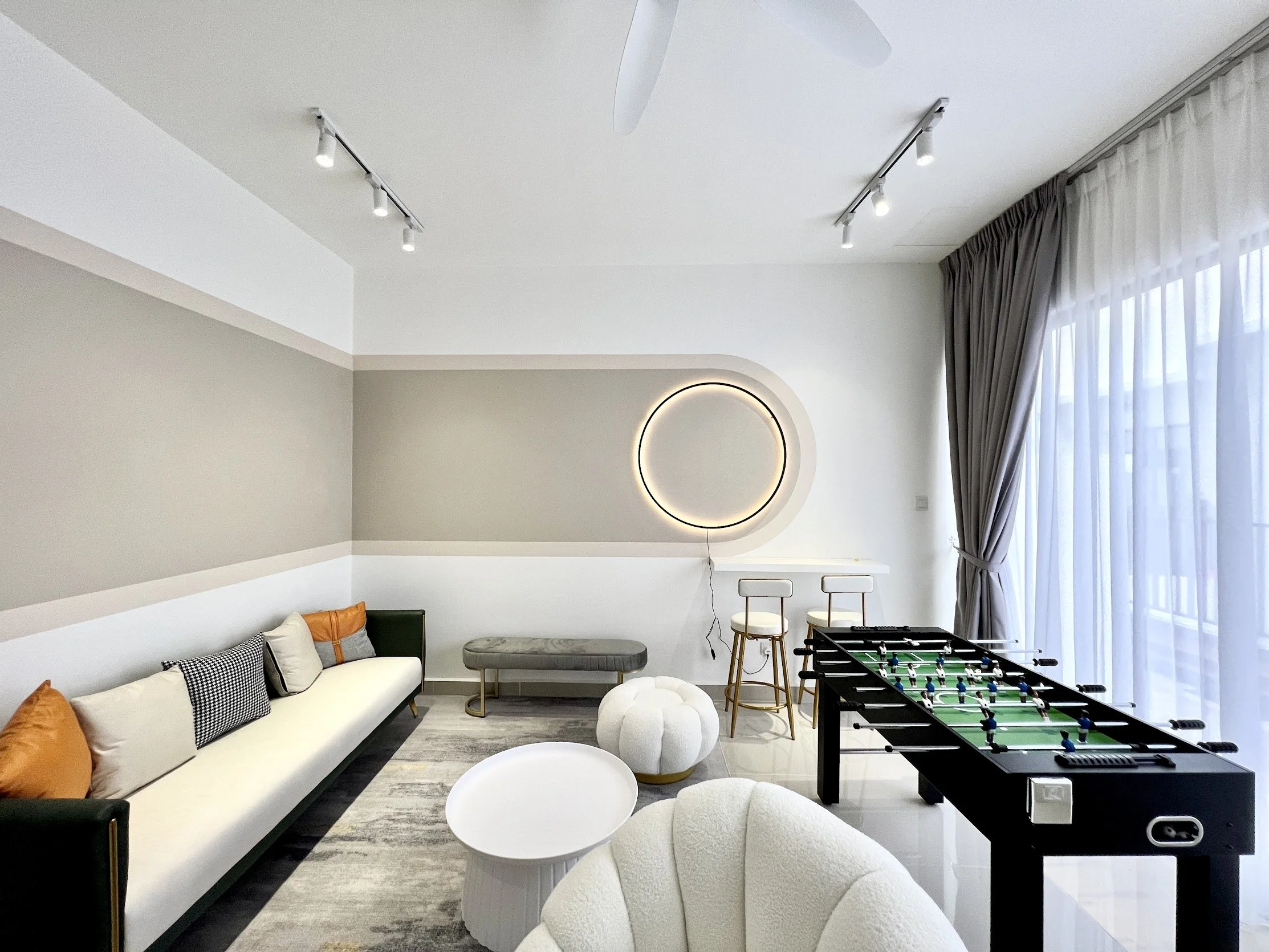 Living room with white sofa, patterned pillows, ottoman, bar stools, foosball table, circular wall light, curtains, and track lighting.