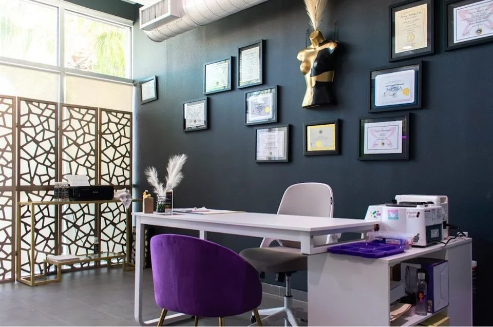 Office with a dark wall decorated with framed certificates and a mannequin bust with a feathered headpiece. There is a white desk, purple chair, and a decorative room divider. On the desk, there are pampas grass in a vase and office supplies.
