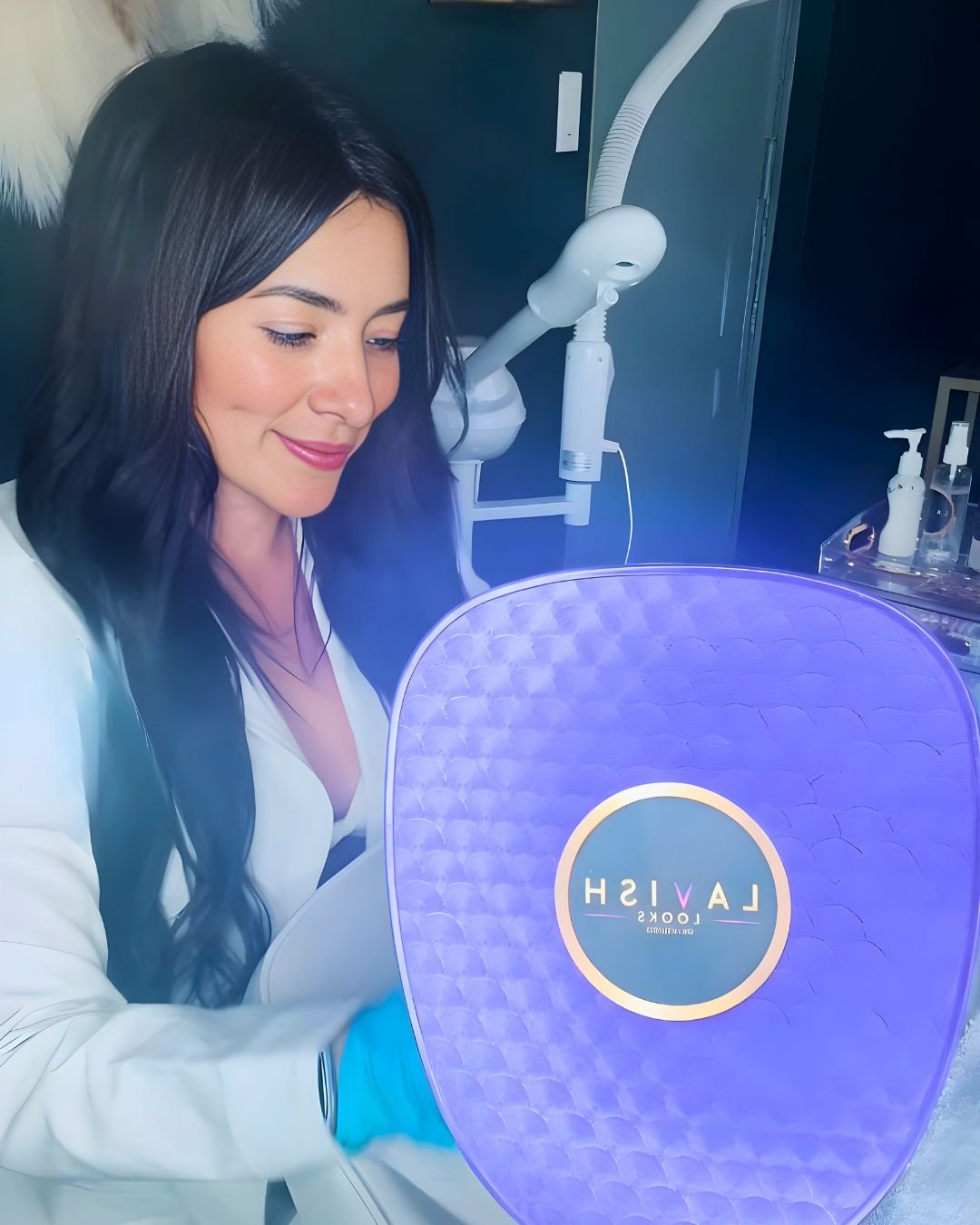 A woman with long dark hair in a white coat smiling while looking through a purple lighted device, with medical equipment and supplies in the background.