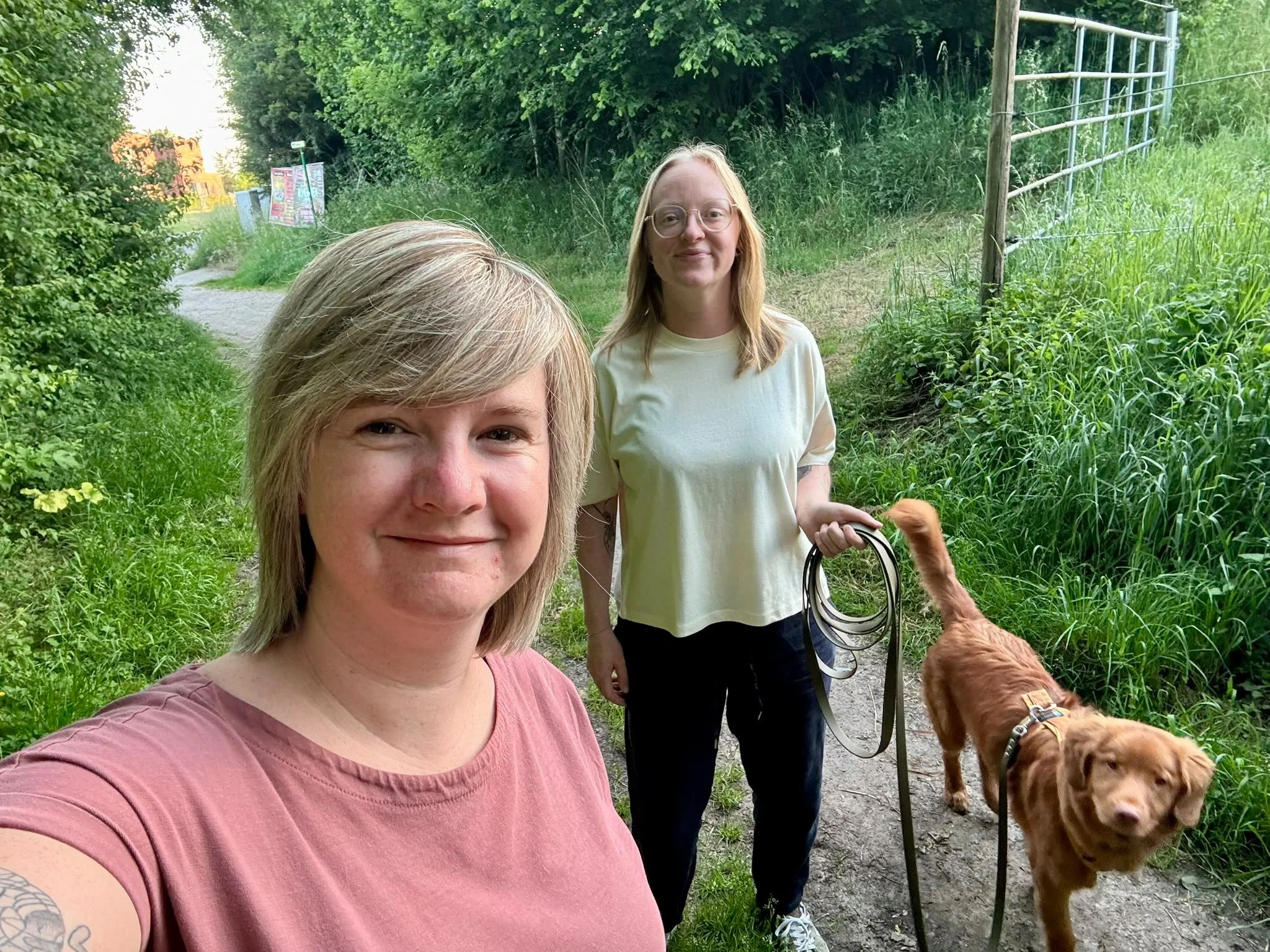 Twee vrouwen wandelen met honden in een groene bosomgeving. Eén vrouw neemt een selfie.