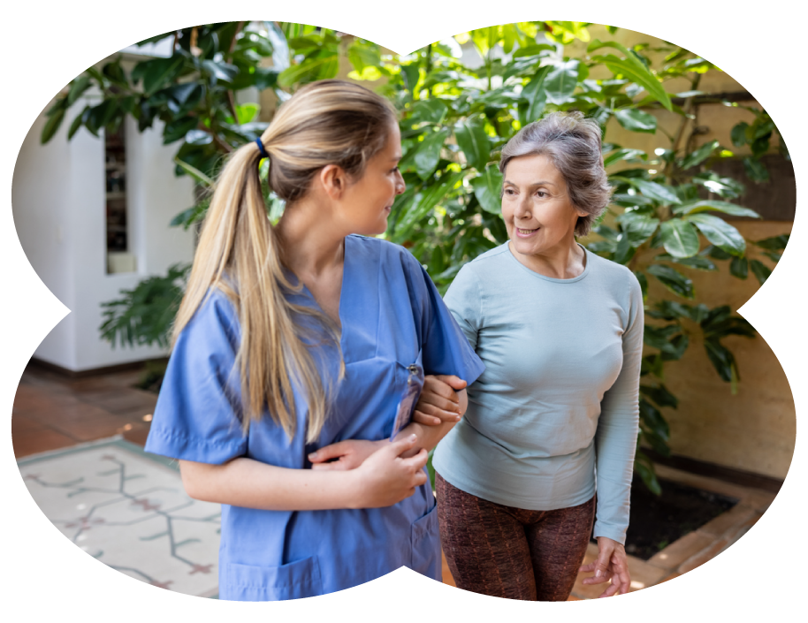 A young female healthcare worker in blue scrubs supporting an older woman with gray hair, both smiling and talking in a lush, green indoor garden.