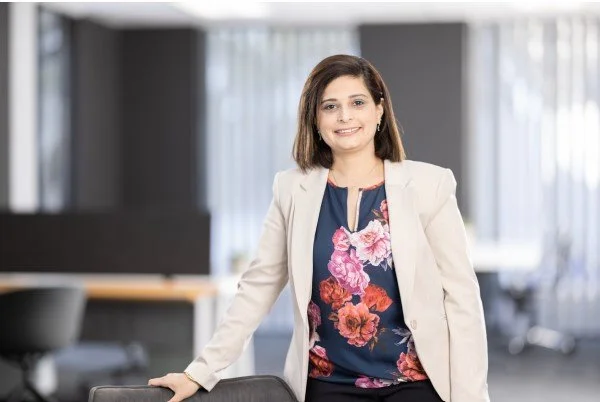 A woman with shoulder-length brown hair, wearing a white blazer over a floral blouse, smiling and standing in an office environment.