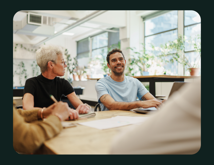 People in a meeting room with large windows and potted plants, engaged in conversation, smiling, with laptops and notebooks on the table.