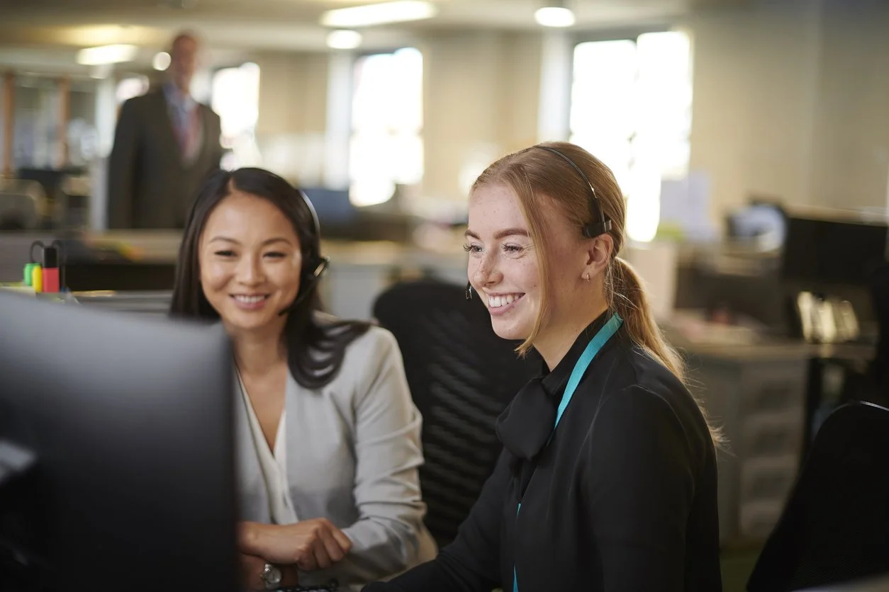 Two women working at a computer in a call center, both wearing headsets and smiling, in a busy office environment.
