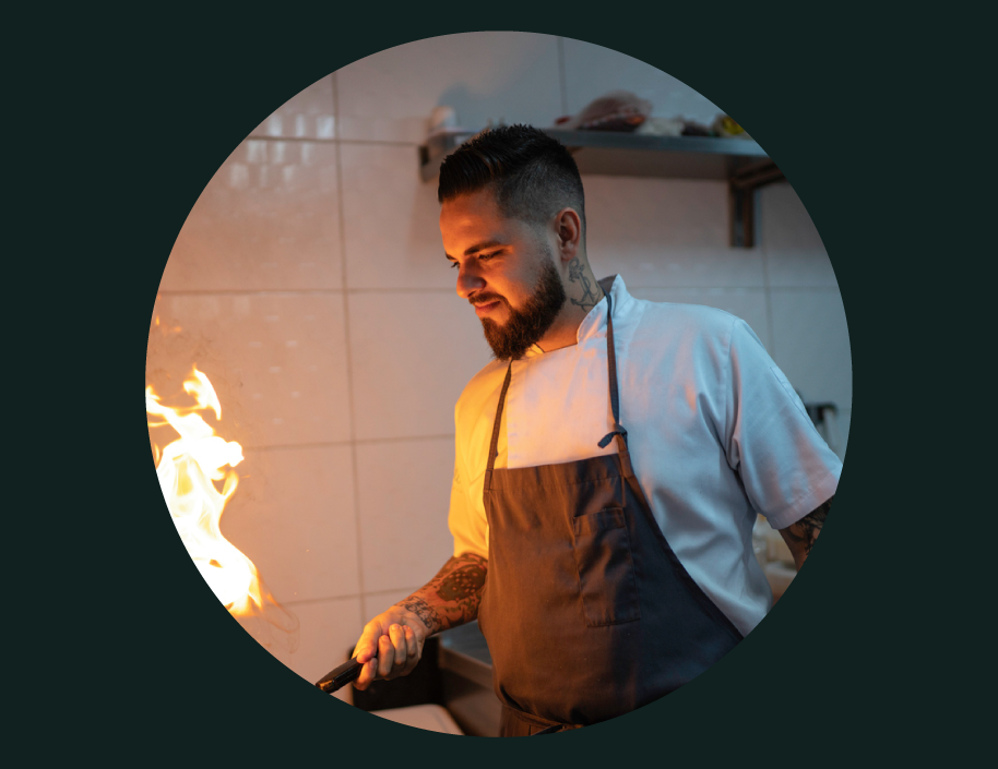 A chef with tattoos and a beard cooking in a kitchen with flames.