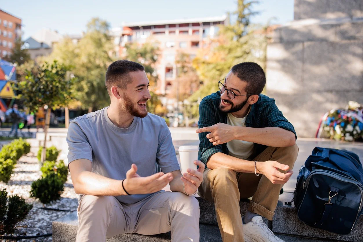 Two young men sitting on a park bench, smiling and chatting in an outdoor urban setting. One is holding a coffee cup, and they seem to be enjoying each other's company.