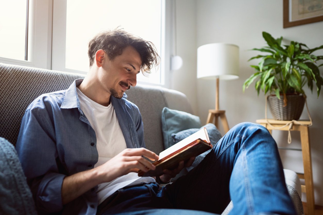 A young man with dark hair and a beard sitting on a sofa, reading a book in a well-lit living room with a potted plant, a lamp, and a framed picture on the wall.