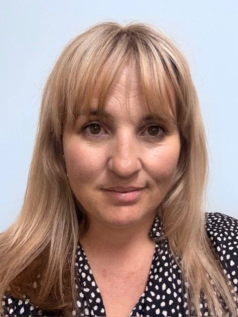 A woman with shoulder-length blonde hair, wearing a black and white polka dot top, smiling softly, against a light blue background.