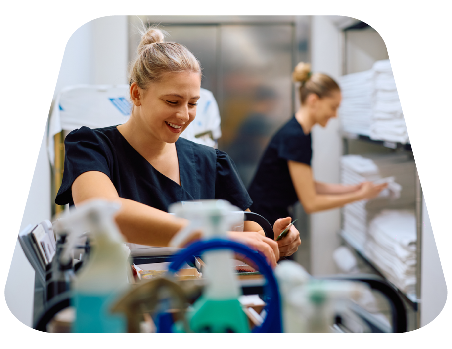 Two women in navy scrubs working in a medical supply room, one smiling at a cart and the other organizing on shelves of neatly folded towels.