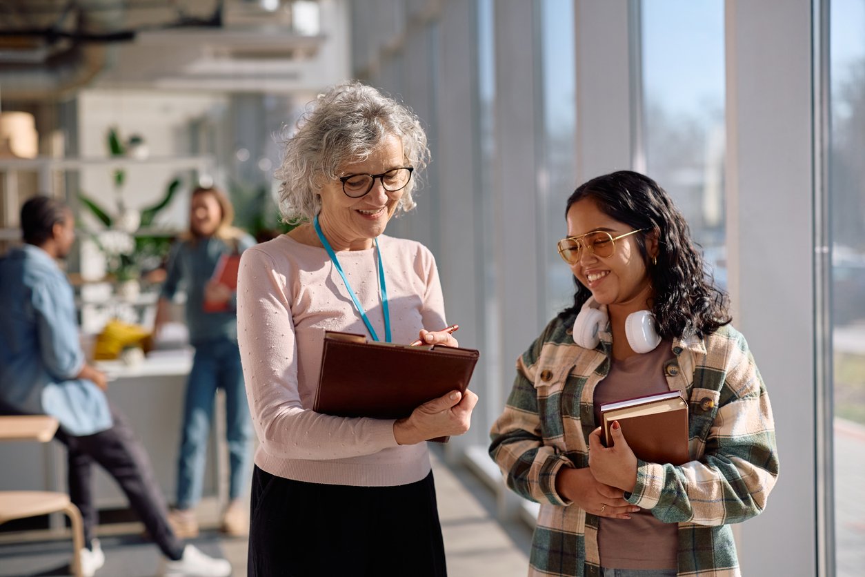 Smiling older woman and younger woman talking in a brightly lit modern indoor space, both holding notebooks, with other people in the background.