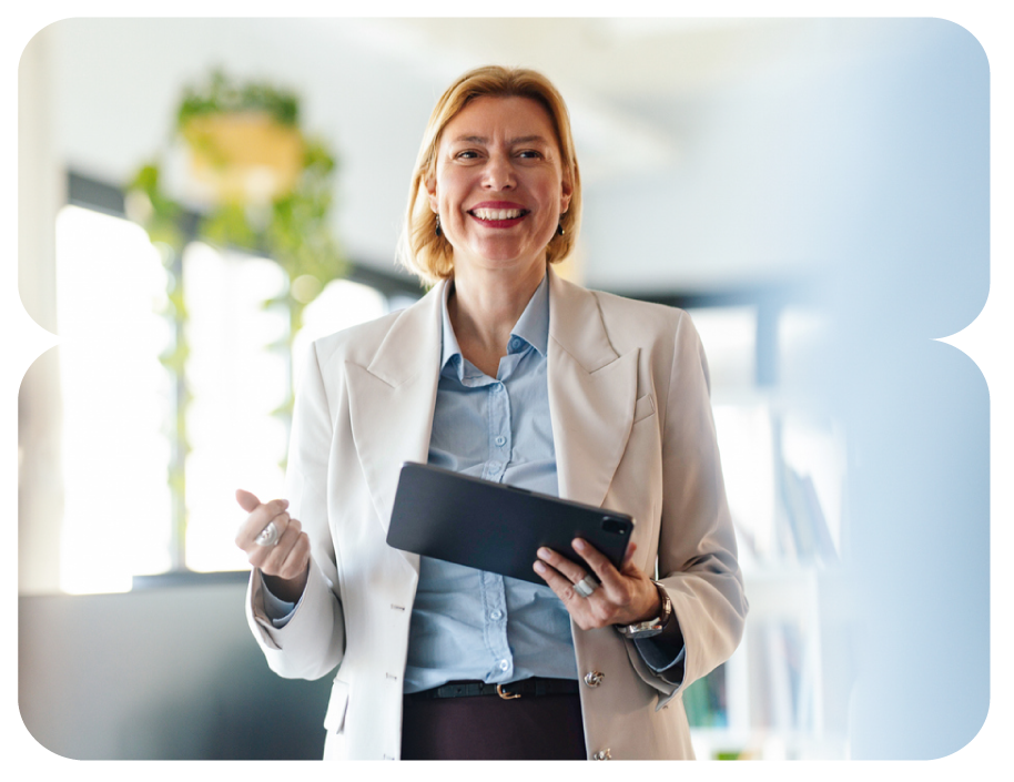 A woman in professional attire smiling while holding a tablet in an office setting.