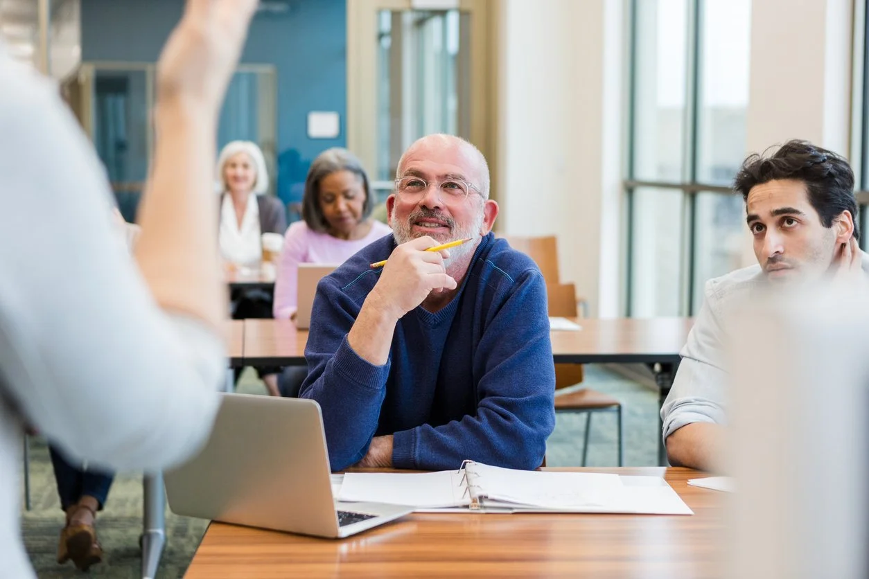 A diverse group of people attending a meeting in a bright conference room, with a man in a blue sweater holding a yellow pencil, listening attentively to a person speaking, who is partially visible.