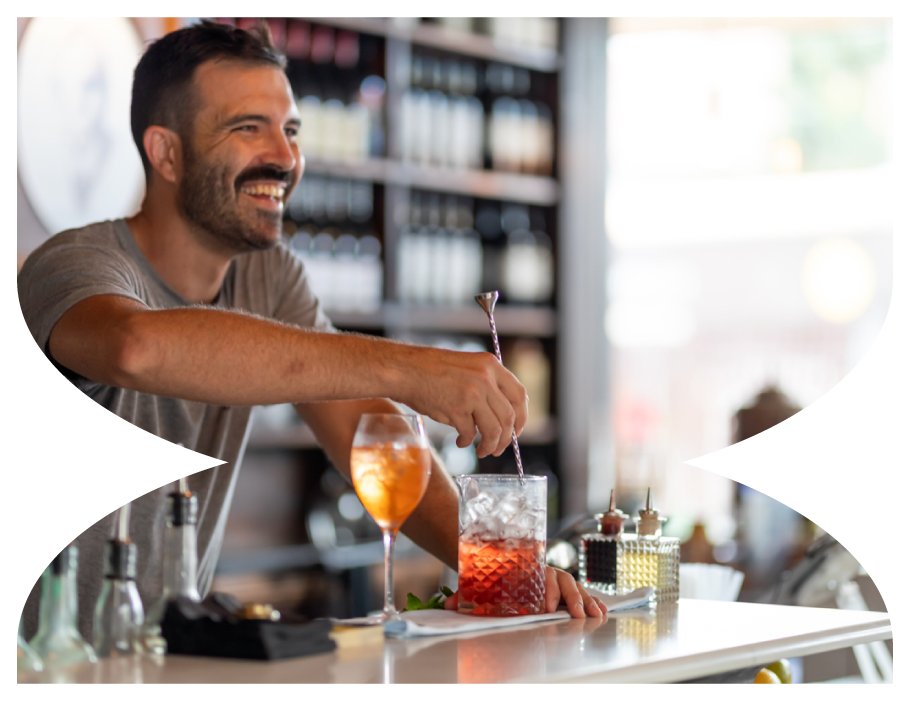 A smiling man with a beard sitting at a bar, stirring a drink in a glass with ice. There are two colorful cocktails on the bar, bottles, and bar tools in the background.