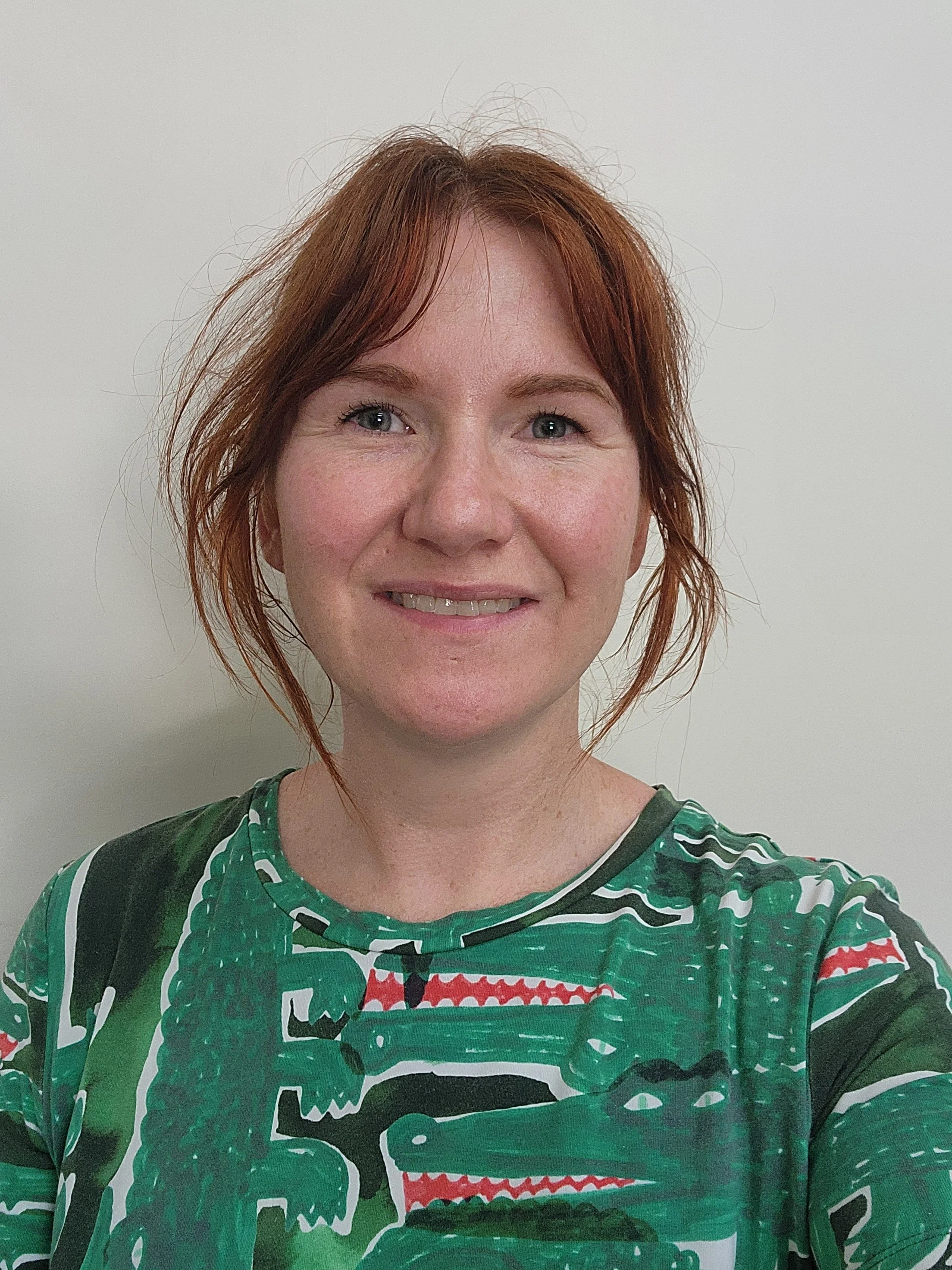 A woman with short, wavy red hair smiling at the camera, wearing a green, black, white, and red patterned sports jersey, standing against a plain white wall.