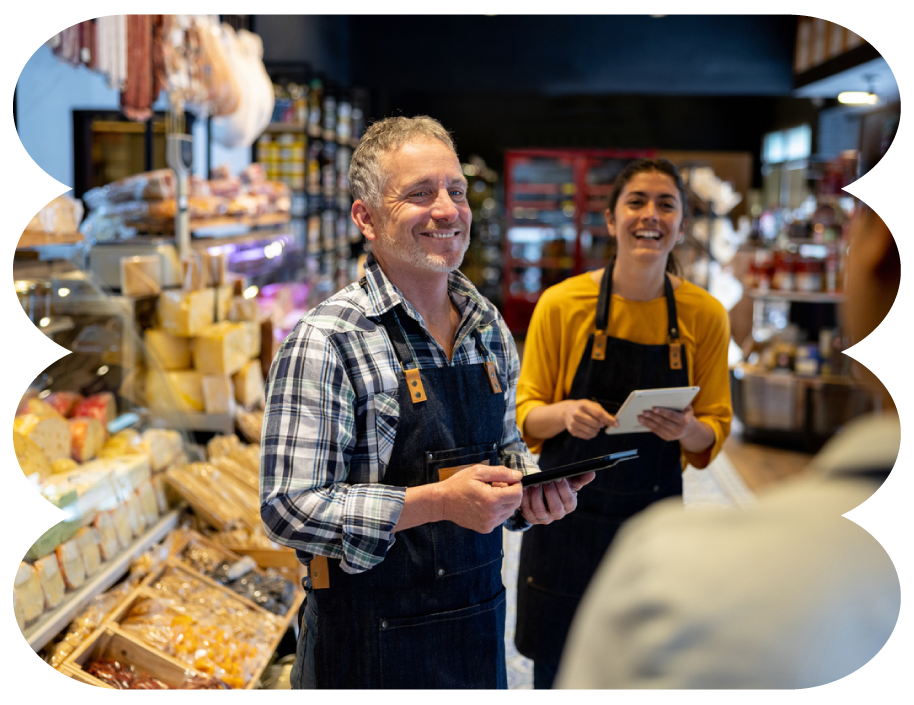 Cheerful middle-aged man and young woman working at a grocery store, smiling and interacting with a customer, surrounded by cheese and other deli products.