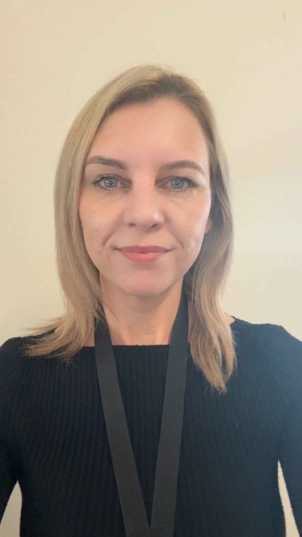 A woman with blonde shoulder-length hair, blue eyes, and a fair complexion, wearing a black top and a black lanyard, standing against a plain light-colored wall.